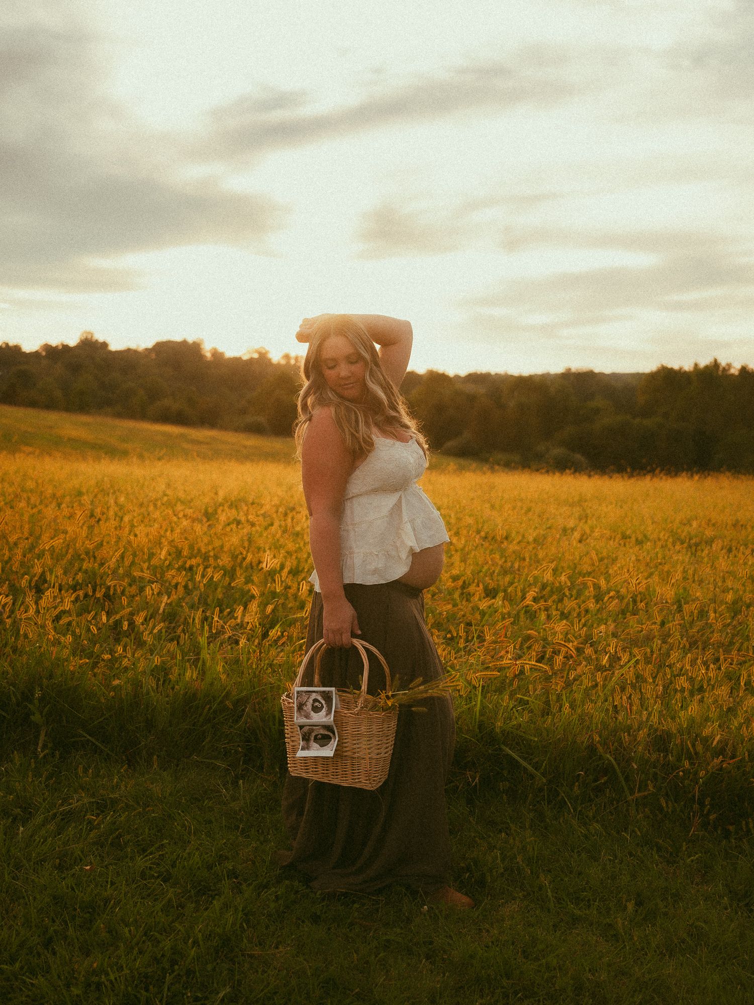 Pregnant woman in white top holds wicker basket in golden sunlit field at sunset, smiling over shoulder.