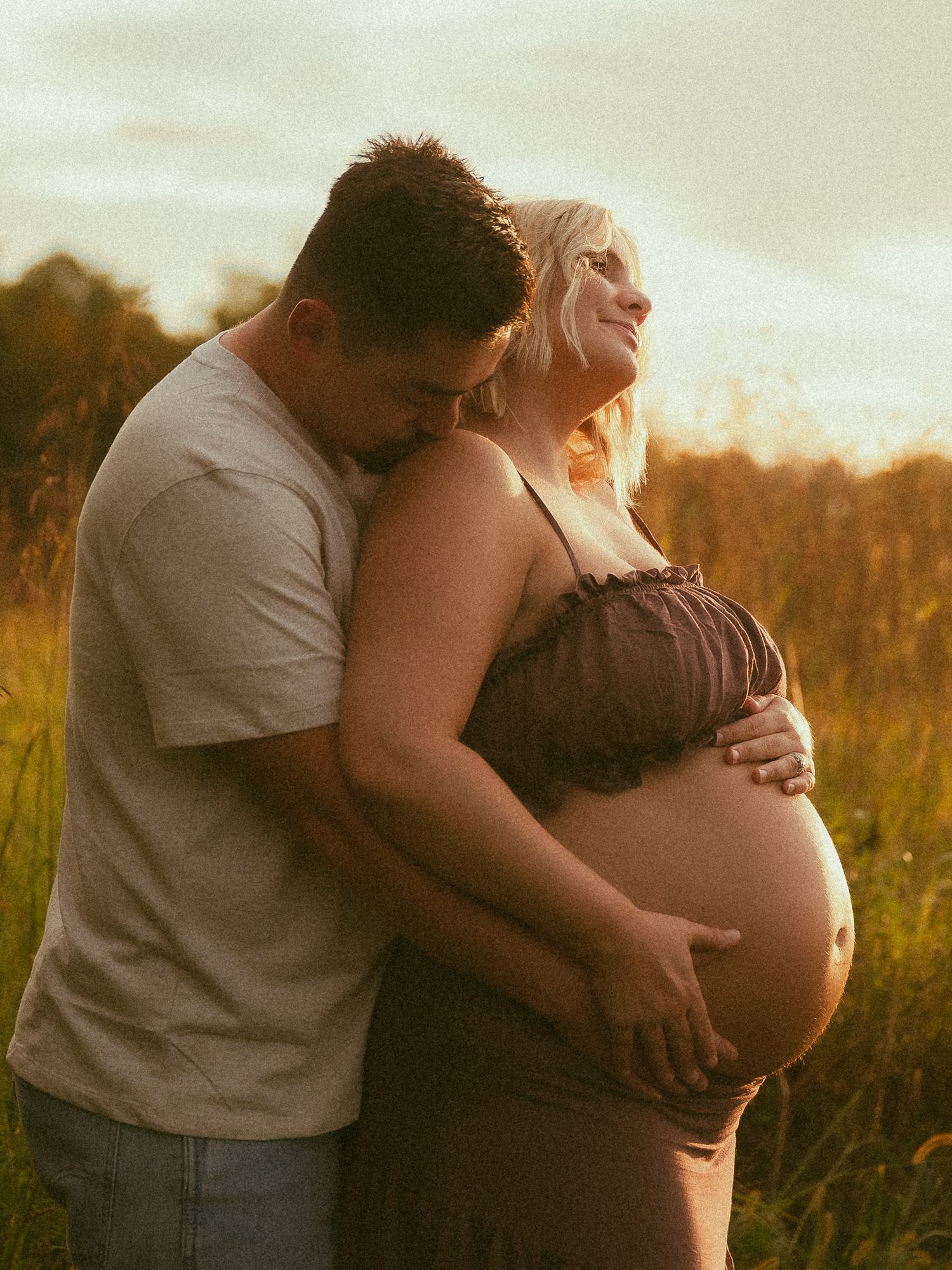 Couple sharing a tender maternity moment in a golden sunset field, man embracing pregnant woman from behind.