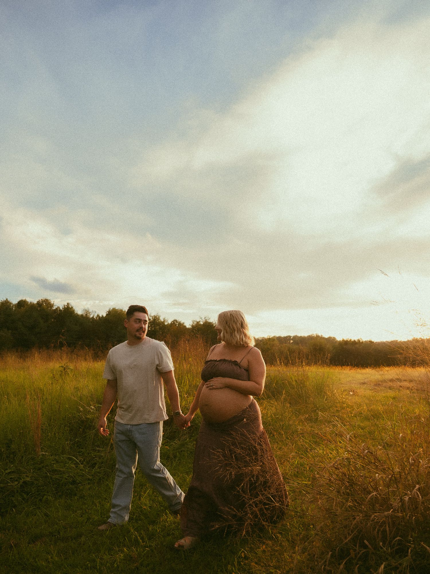 Couple holding hands in golden field at sunset, pregnant woman in brown dress showing baby bump, maternity photo.