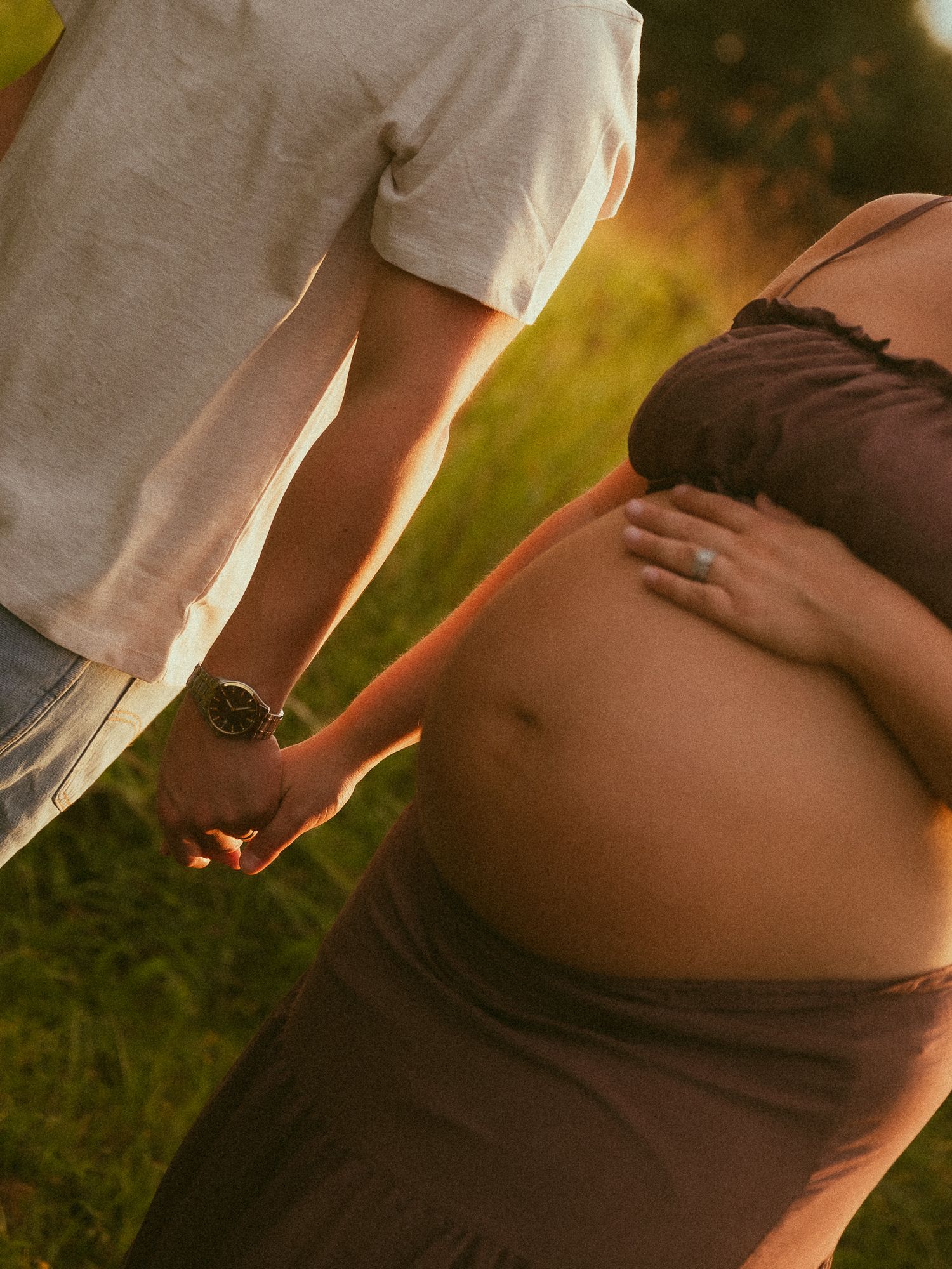 Close-up of pregnant woman cradling baby bump with partner holding hands outdoors in warm sunset light.