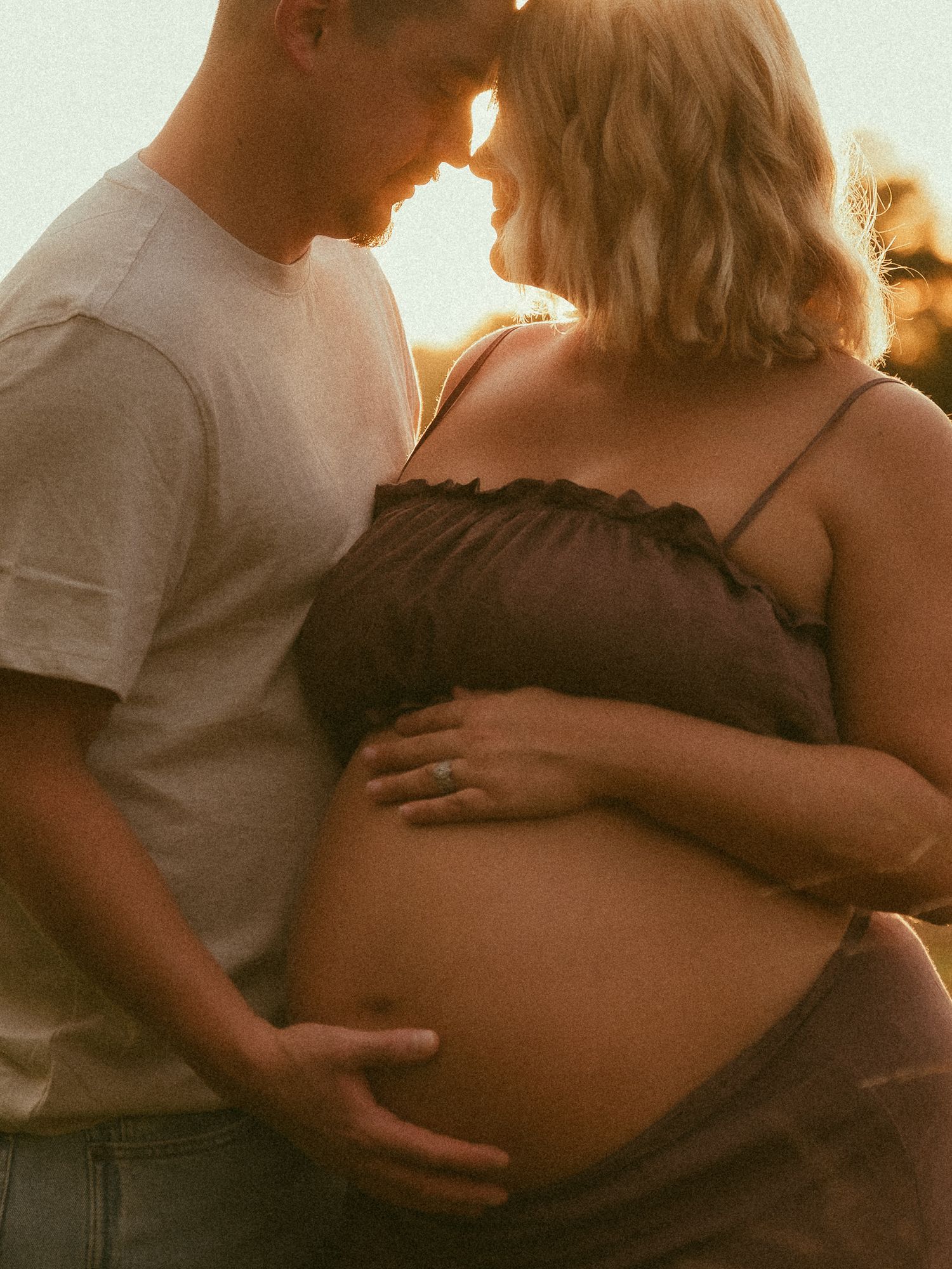 Couple sharing intimate maternity moment outdoors at golden hour, man embracing pregnant woman's belly in warm sunlight.