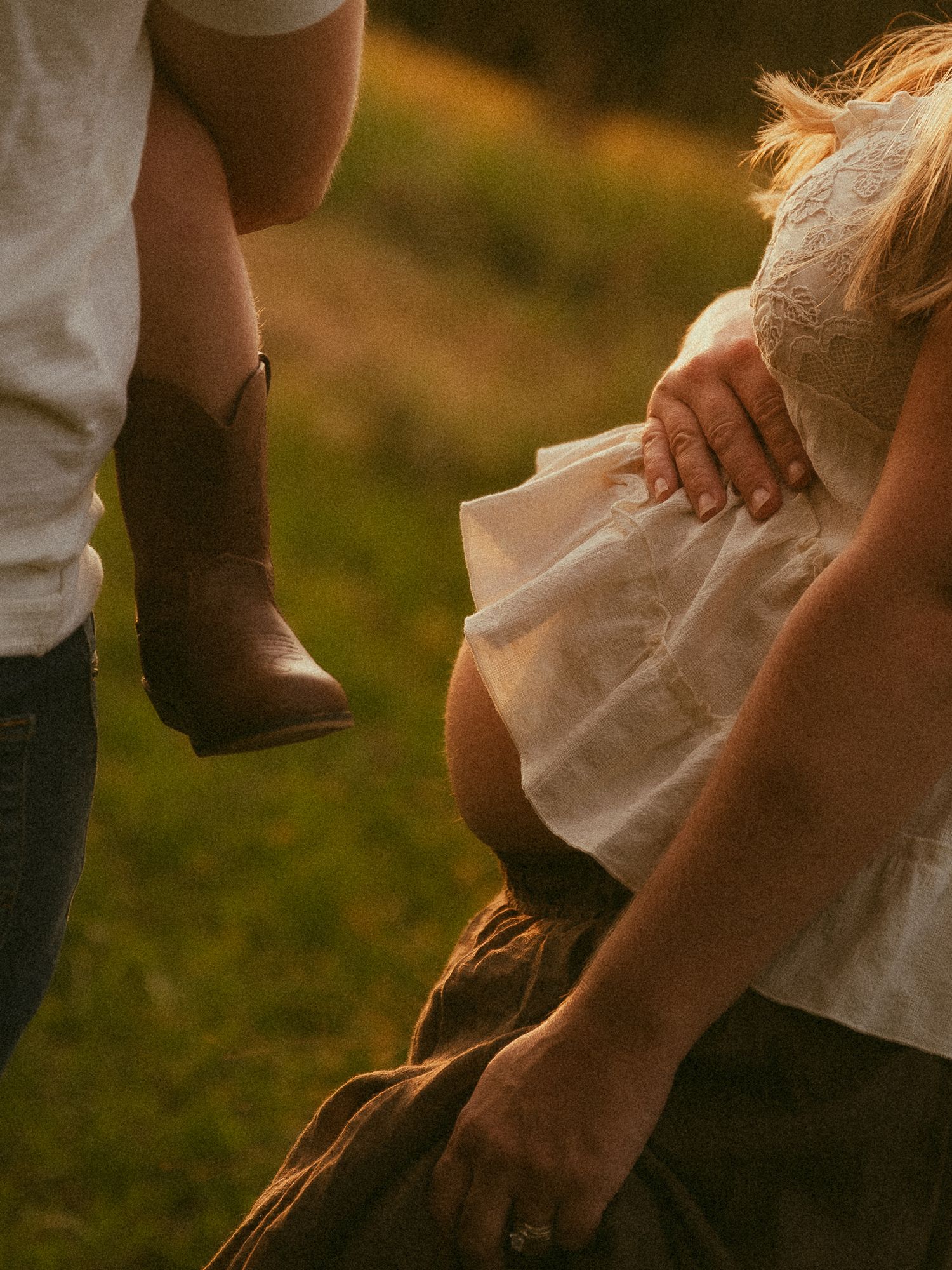 Golden hour maternity photo of pregnant woman in white dress with partner wearing cowboy boots outdoors.