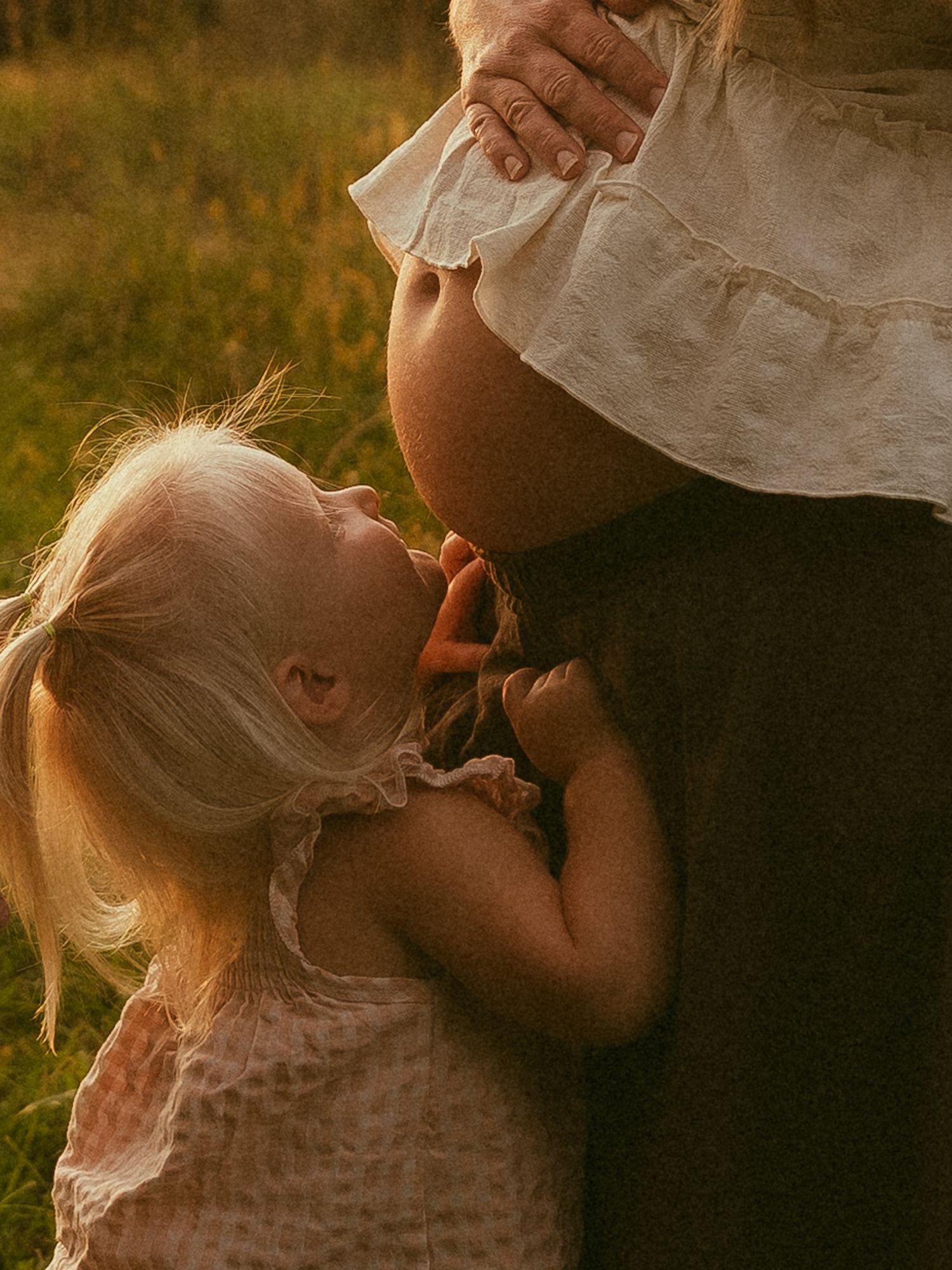 A young blonde toddler girl kisses a pregnant mother's bare belly in a golden sunlit field.