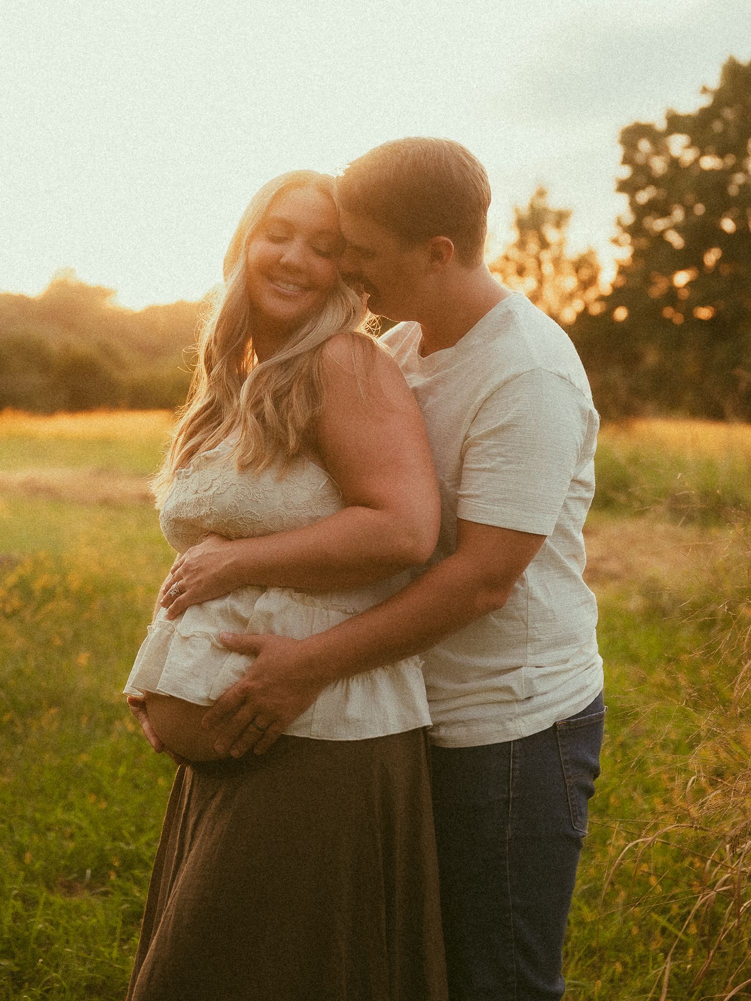 A pregnant woman in white lace and olive skirt embraces her partner in a sunlit meadow.