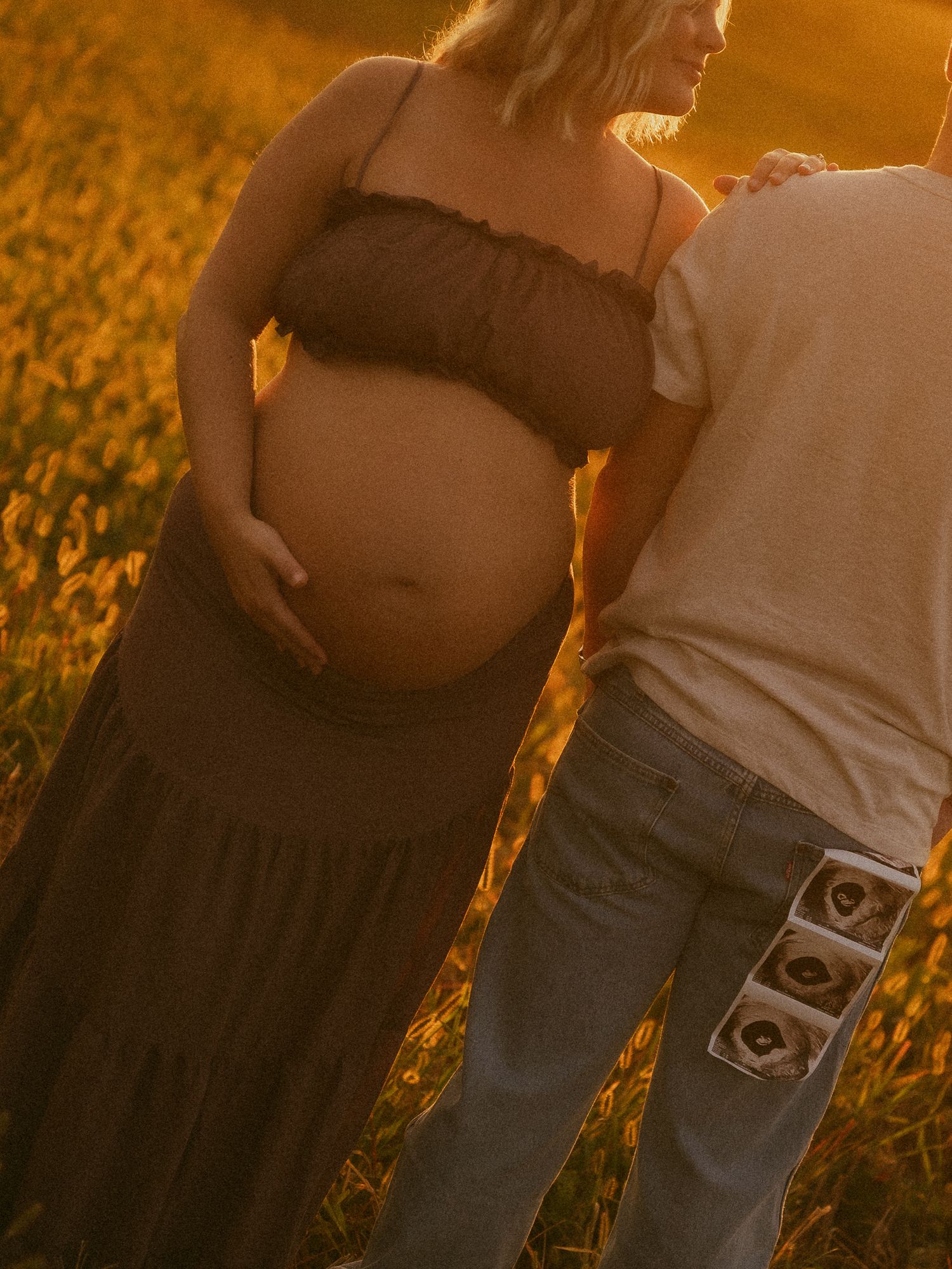 Pregnant woman in black crop top and maxi skirt holds baby bump at golden sunset while partner holds ultrasound photos.