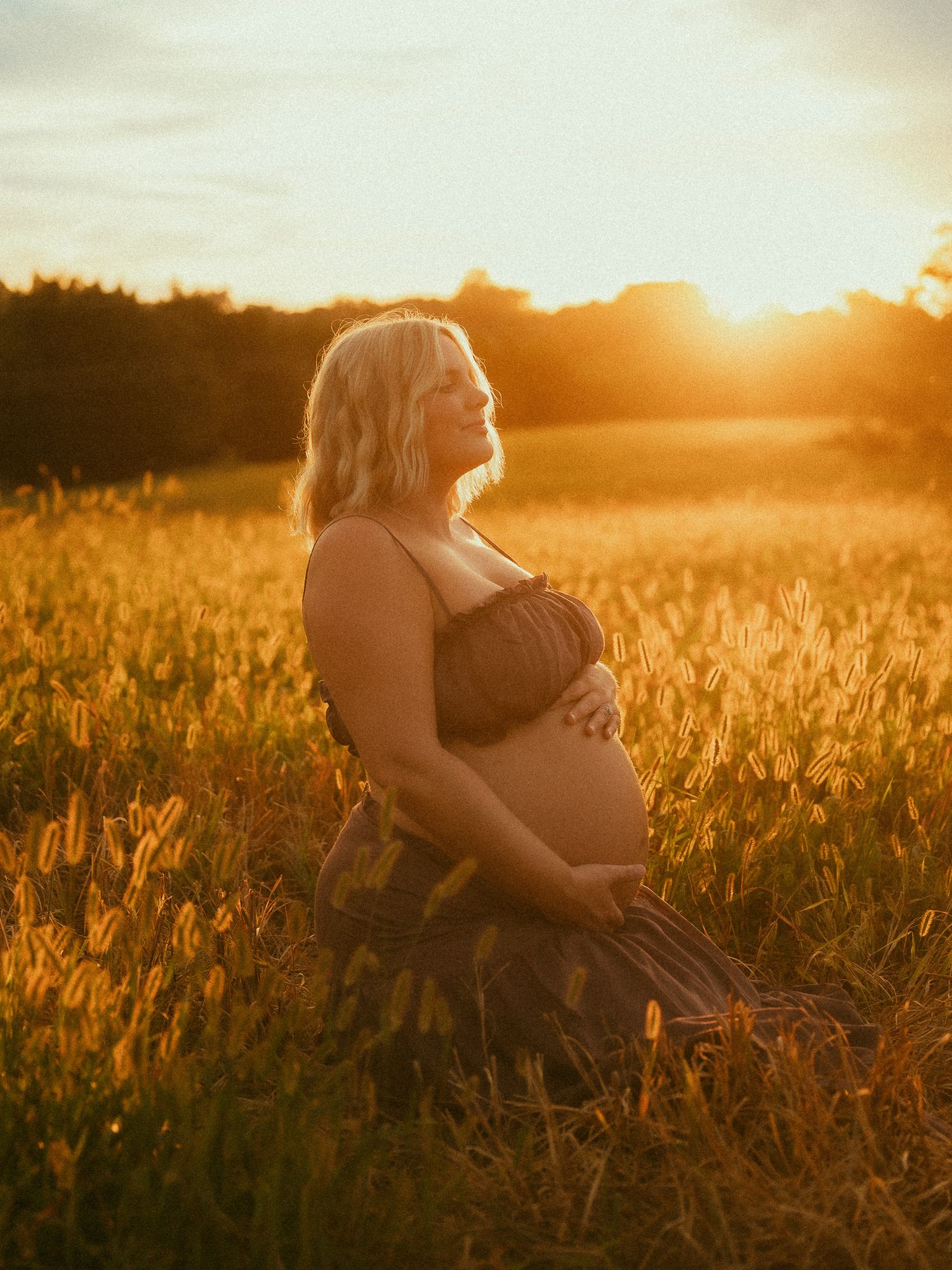 Pregnant woman in dark dress kneeling in golden field at sunset, cradling baby bump while gazing upward.