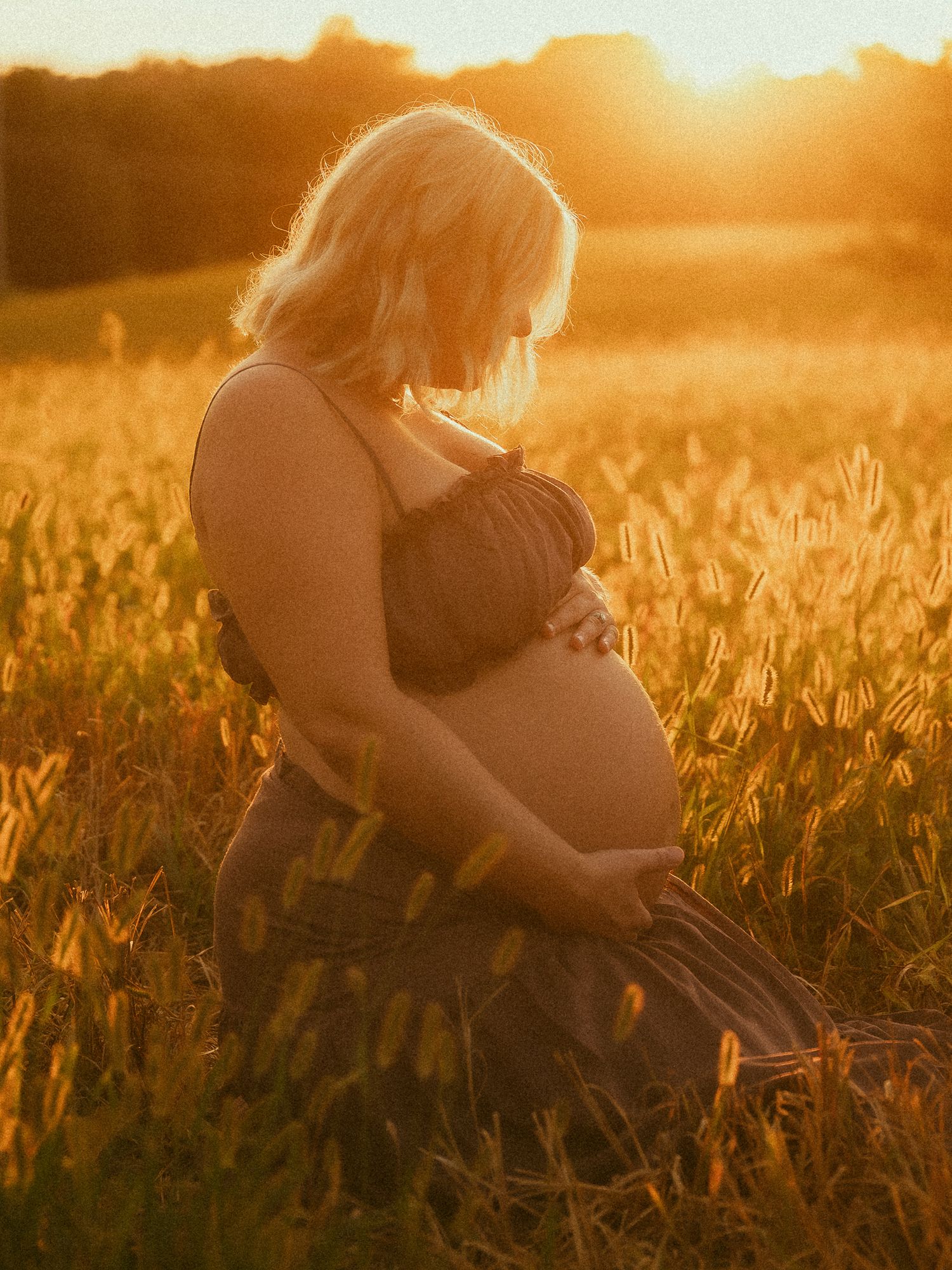 Pregnant woman in dark dress sitting in sunlit field at golden hour, looking down and holding her baby bump.