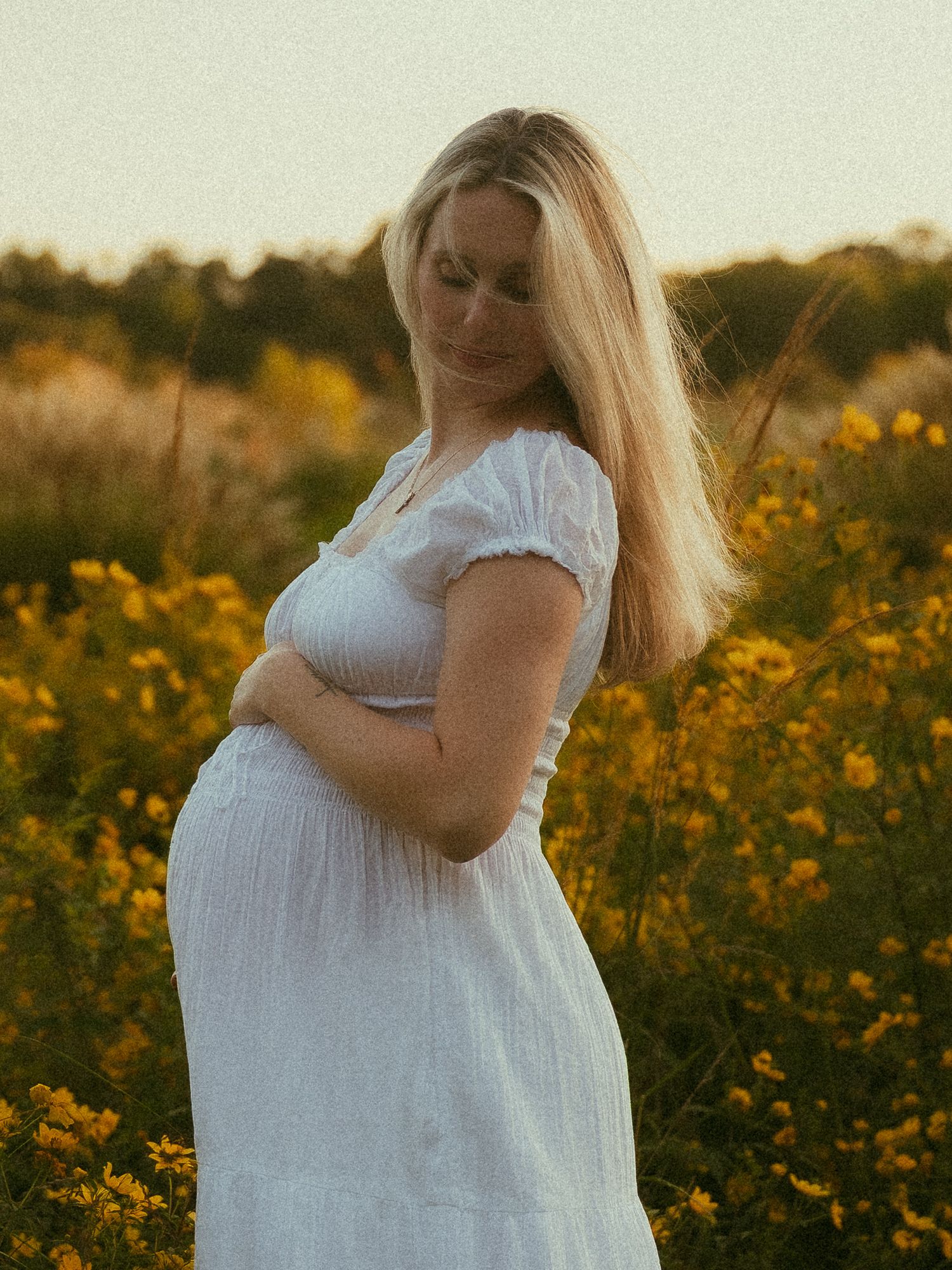 Pregnant woman in white dress cradling baby bump in golden wildflower field at sunset.
