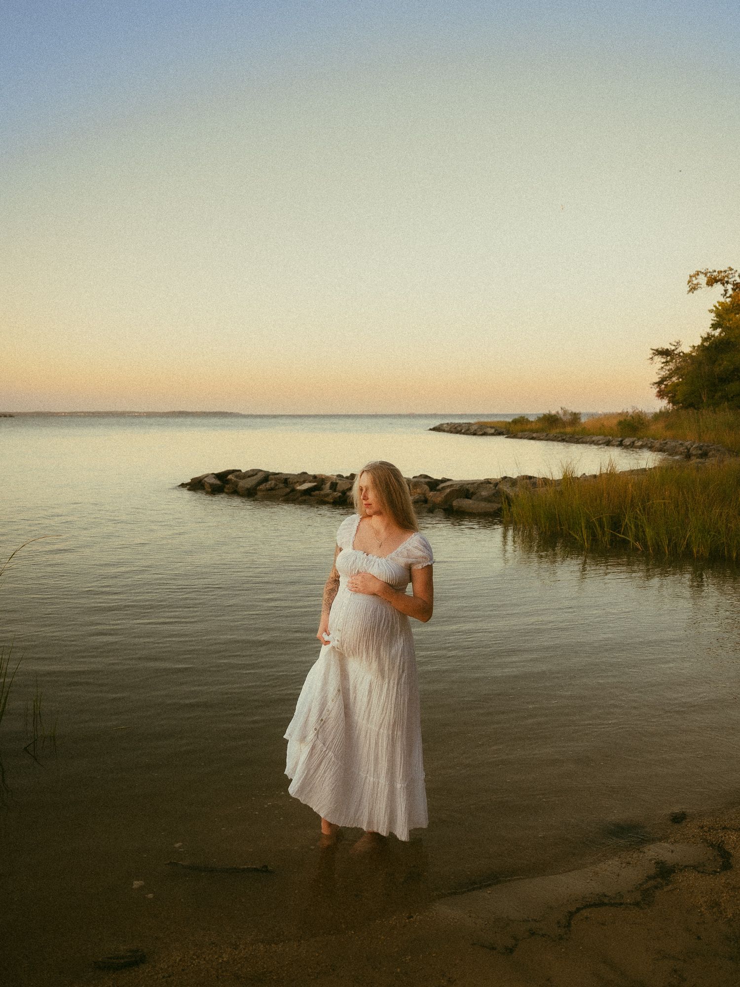 Pregnant woman in white dress standing in shallow water at sunset by a rocky shoreline.