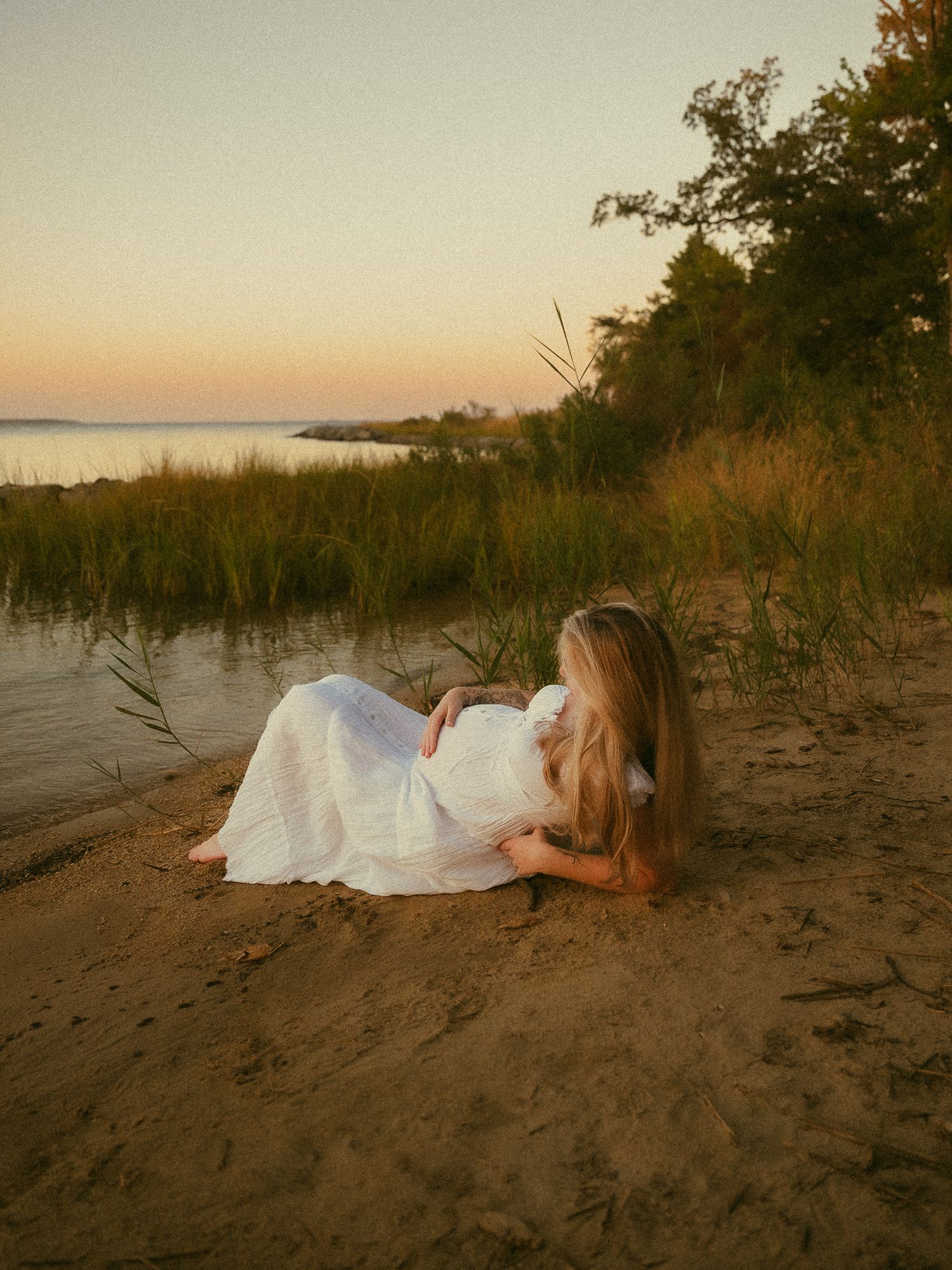 A blonde woman in a white dress reclines on sandy beach at golden sunset near calm water and tall grass.