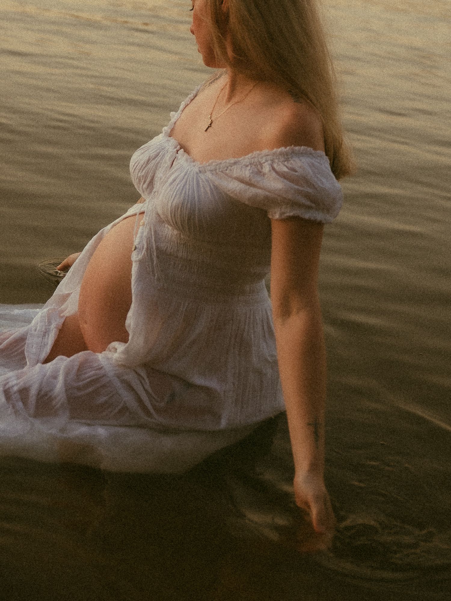 Pregnant woman in white off-shoulder dress sitting in shallow water at golden hour sunset.