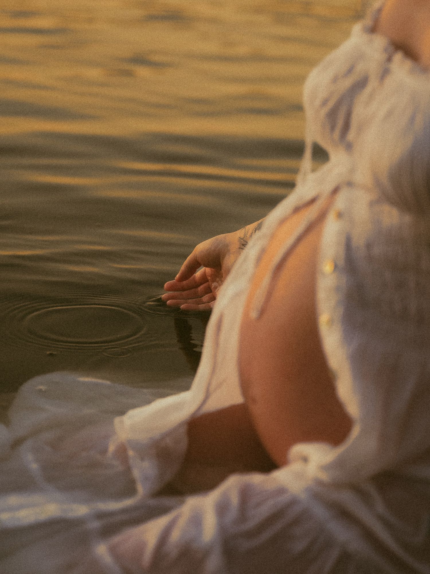 Pregnant woman in white dress sitting by golden water, gently touching the surface during a serene maternity photoshoot.
