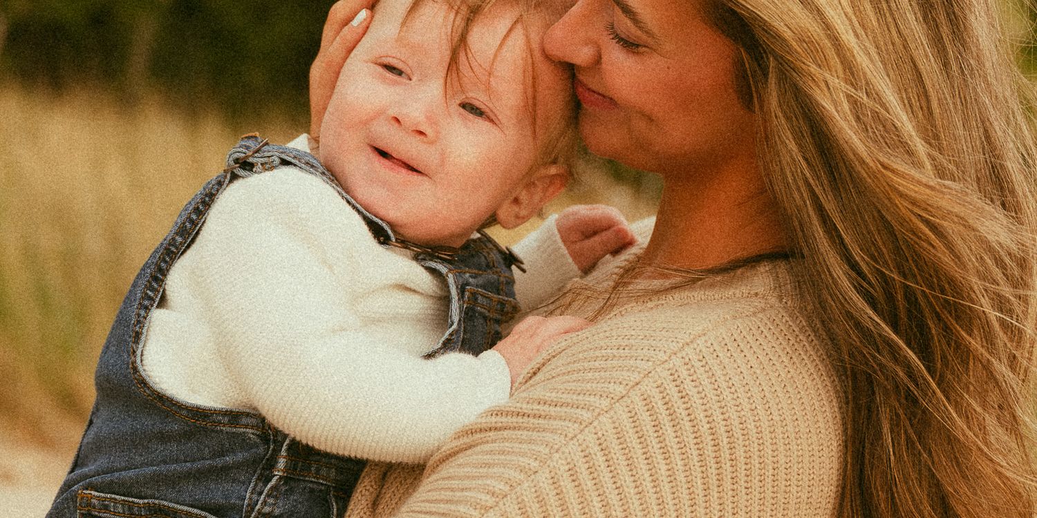 A mother tenderly kisses her smiling baby dressed in denim overalls outdoors in a warm, natural setting.