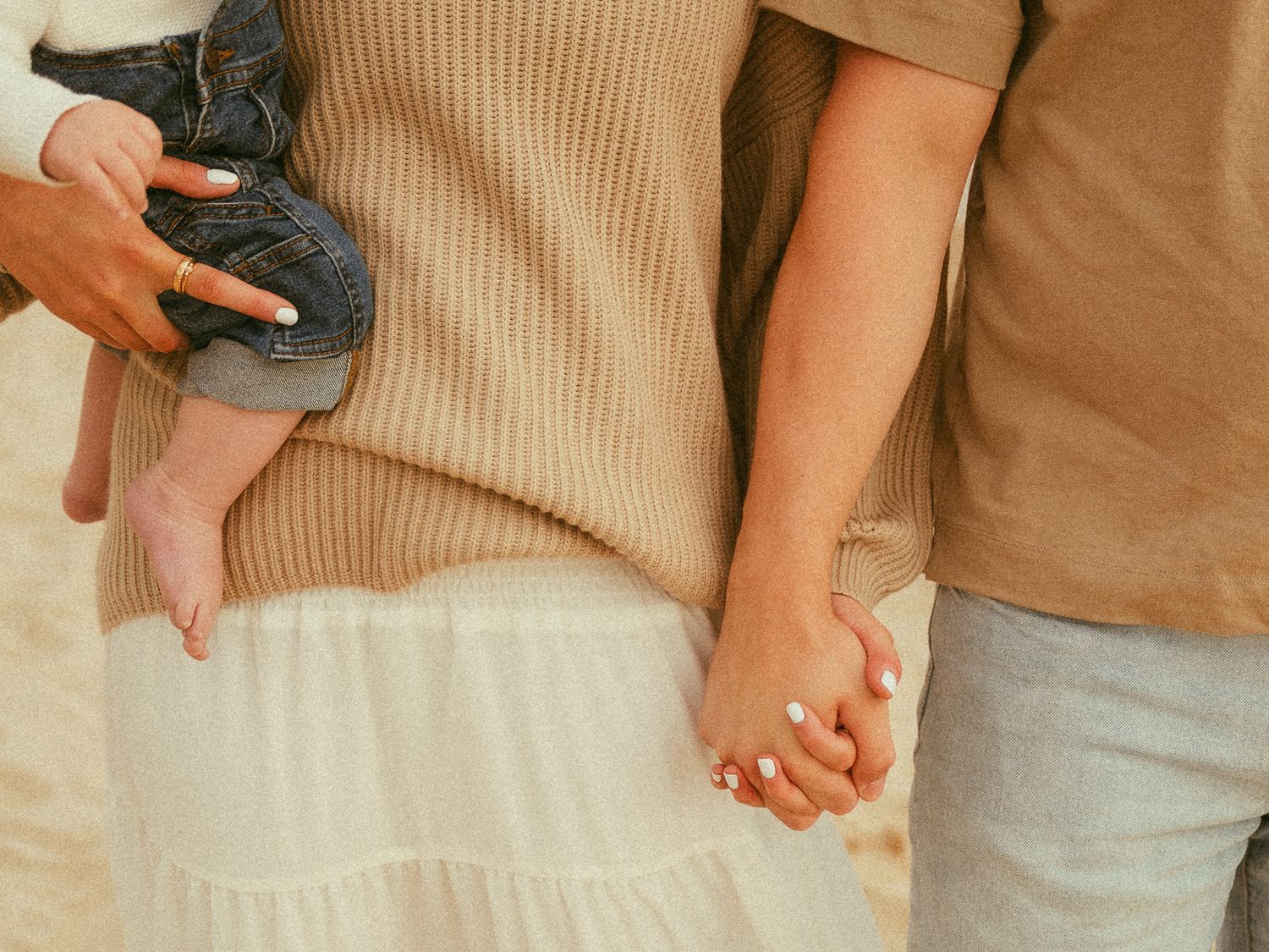 Couple holding hands while carrying a baby, wearing neutral tones, standing outdoors in a warm sandy setting.