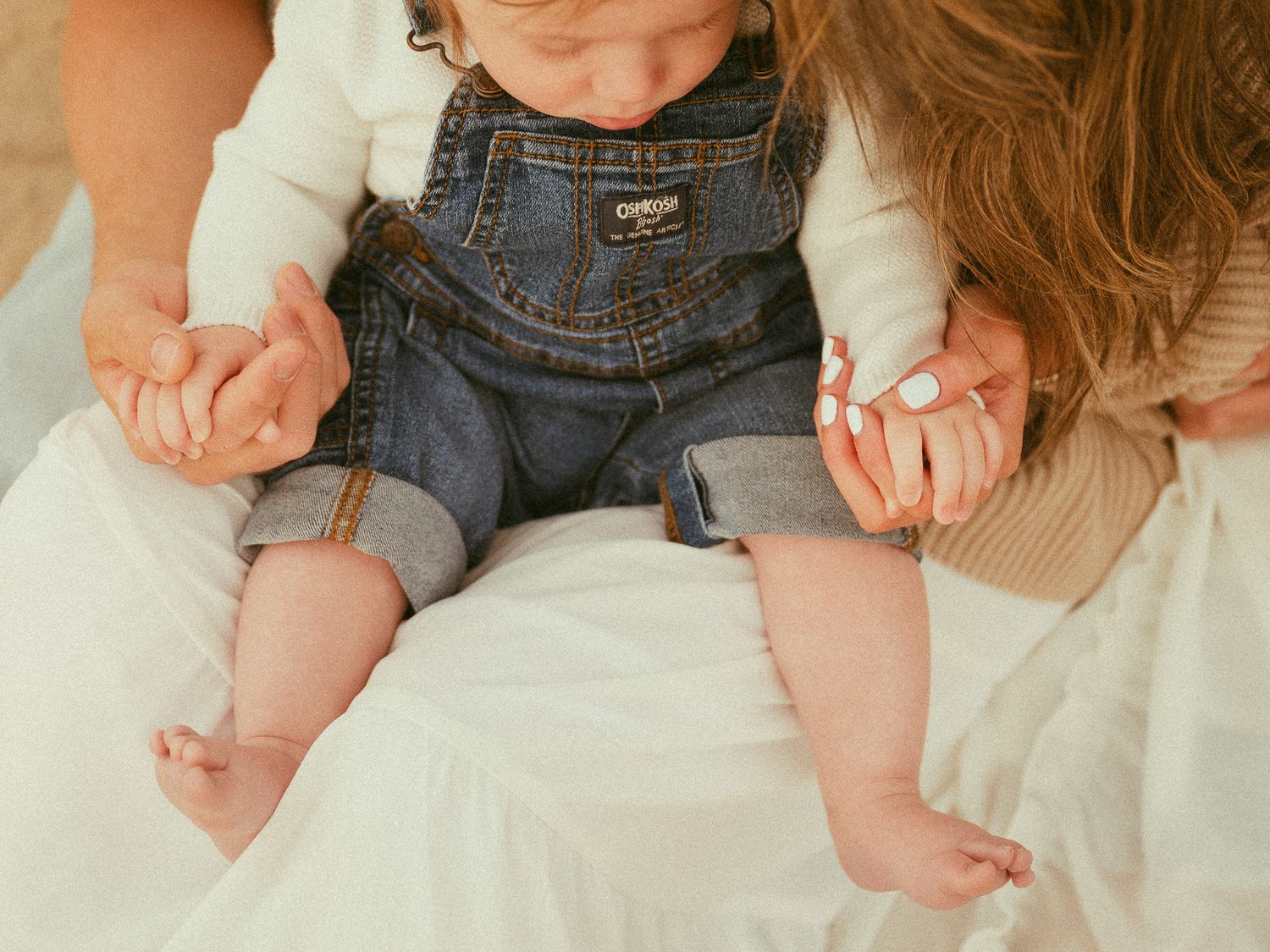 A baby in OshKosh denim overalls sits on a woman's lap while she holds their tiny hands.