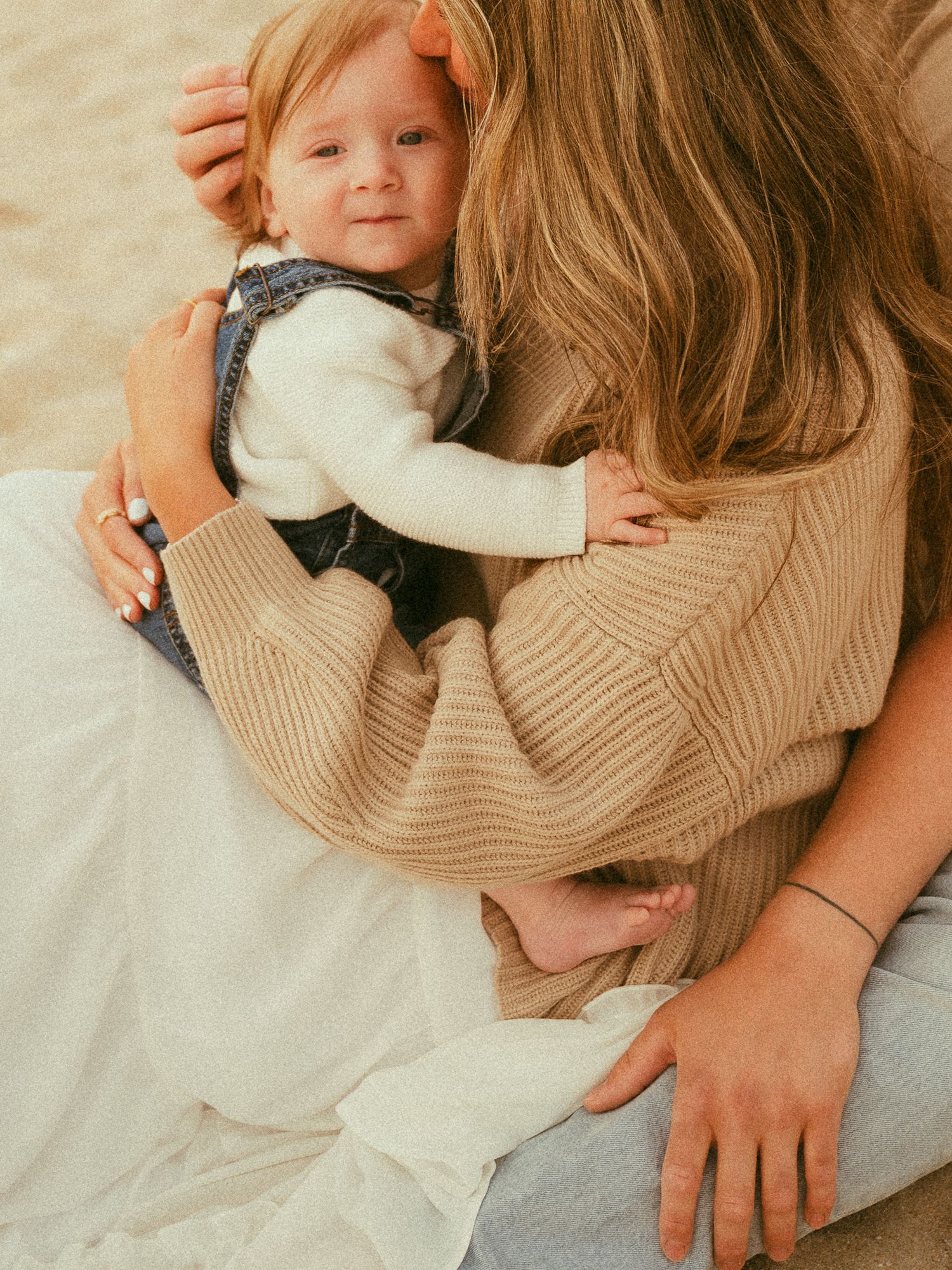 A mother in a beige knit sweater tenderly holds her smiling baby dressed in denim overalls on a sandy beach.