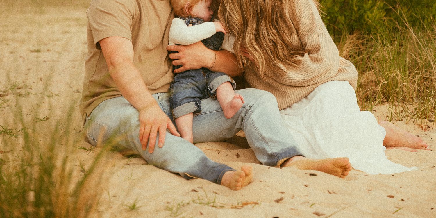 A couple sits on sandy beach holding their baby, wearing casual neutral tones in a warm outdoor family portrait.