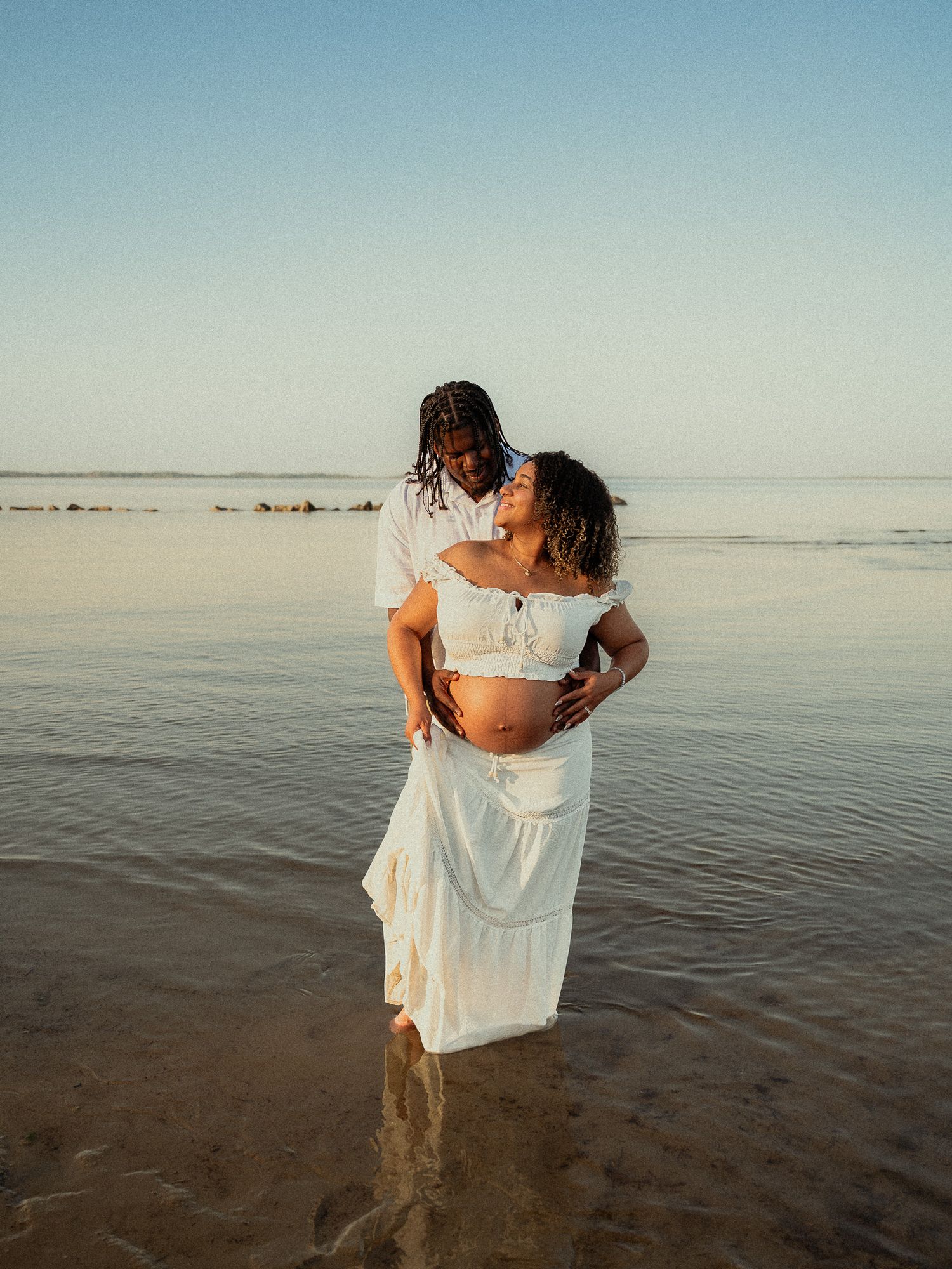 Couple in white outfits embracing in shallow ocean water during a romantic maternity beach photoshoot.