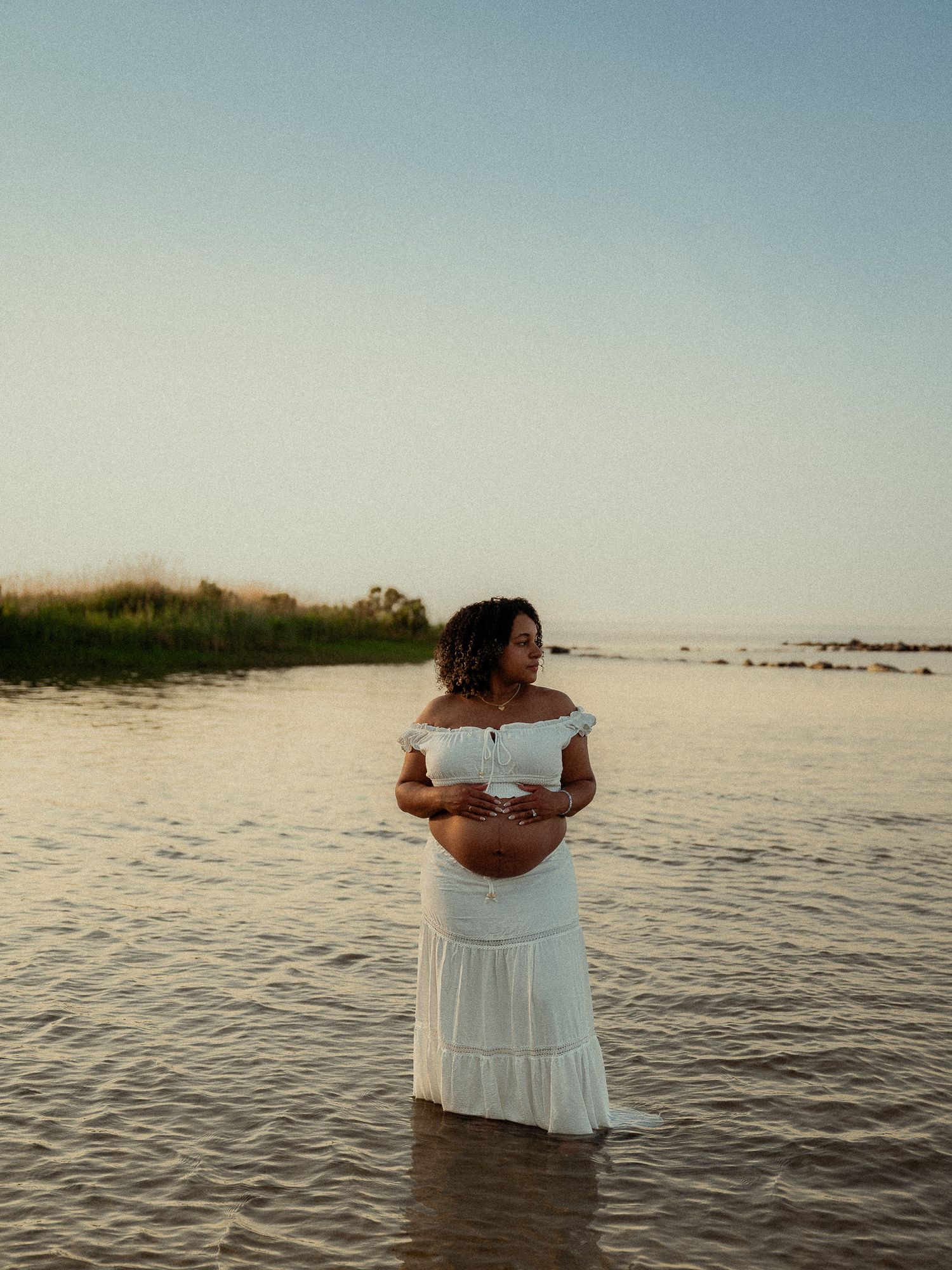 Pregnant woman in white off-shoulder dress standing in shallow water during a serene beach maternity session.