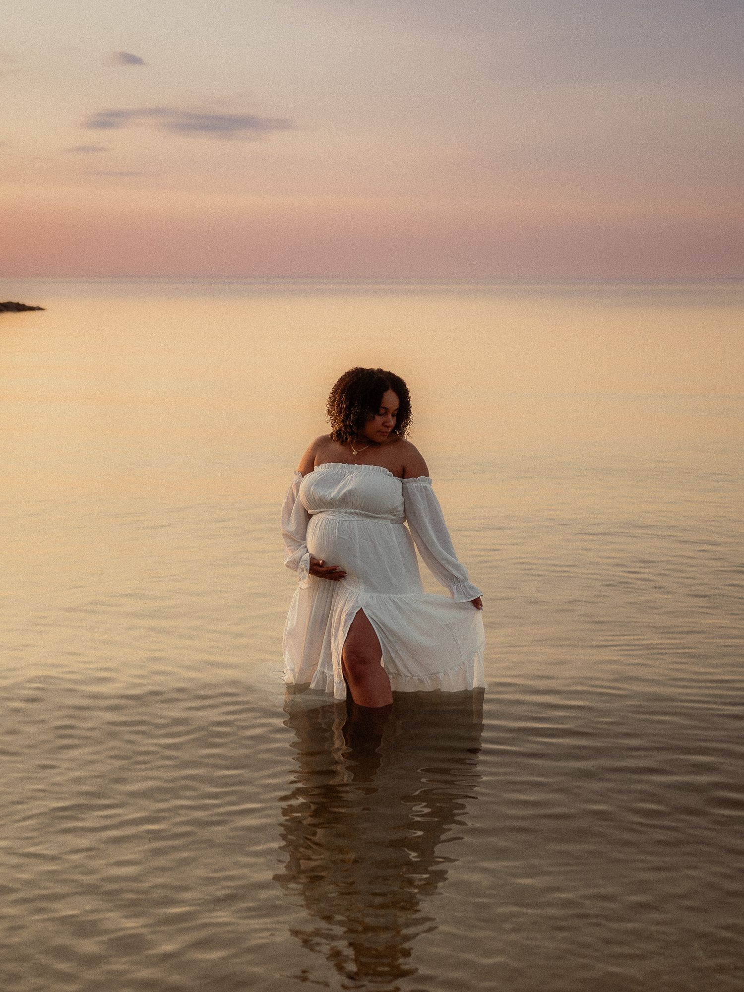 Pregnant woman in flowing white off-shoulder dress standing in calm ocean water at sunset.