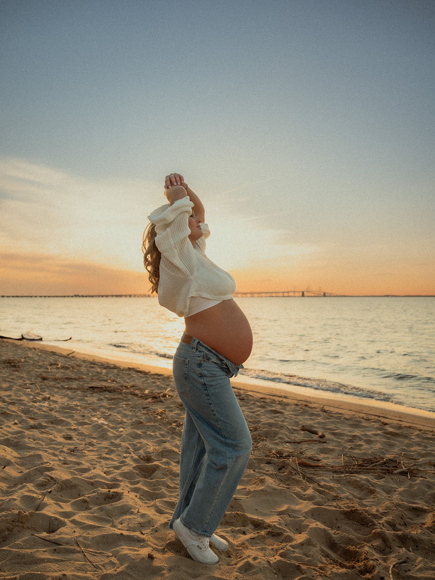 Pregnant woman in white crop top and jeans posing on beach at golden hour sunset.