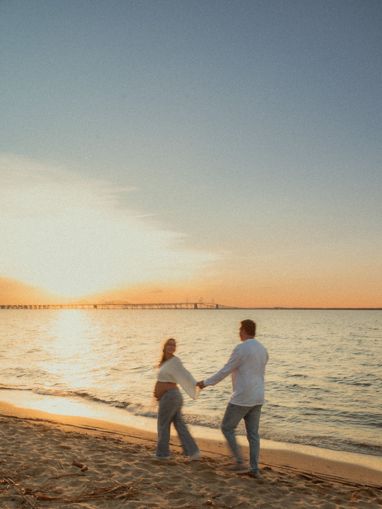 Expecting couple holding hands walking along sandy beach at stunning sunset with pier in background.