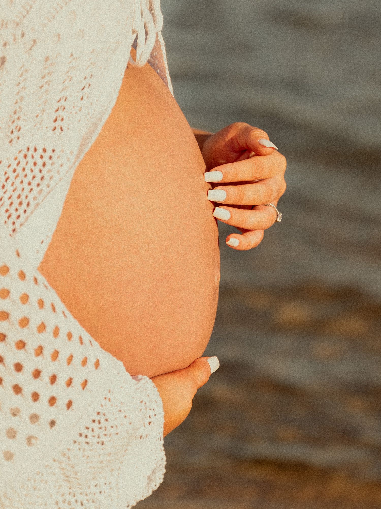 Pregnant woman in white crochet top cradling baby bump near water, showcasing white manicured nails and engagement ring.