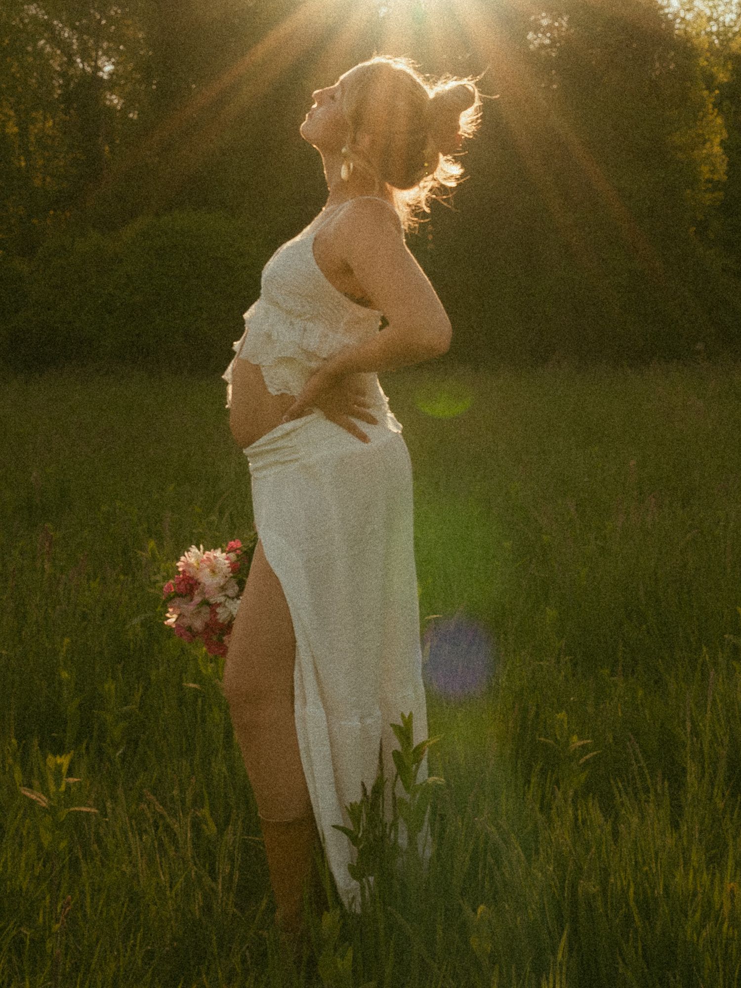 Pregnant woman in white dress holding pink flowers in sunlit meadow during golden hour.