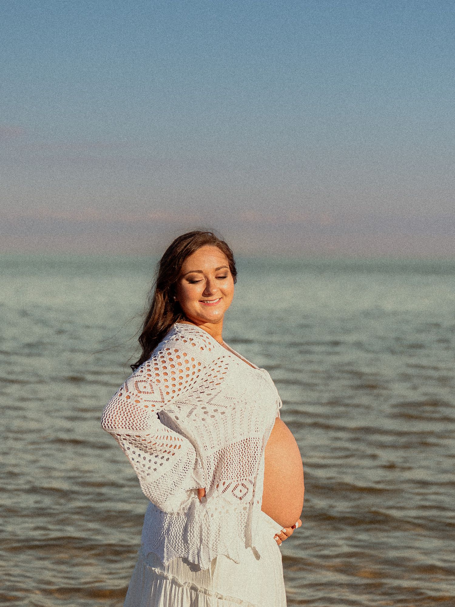Smiling pregnant woman in white crochet cardigan standing by ocean during maternity photo shoot.