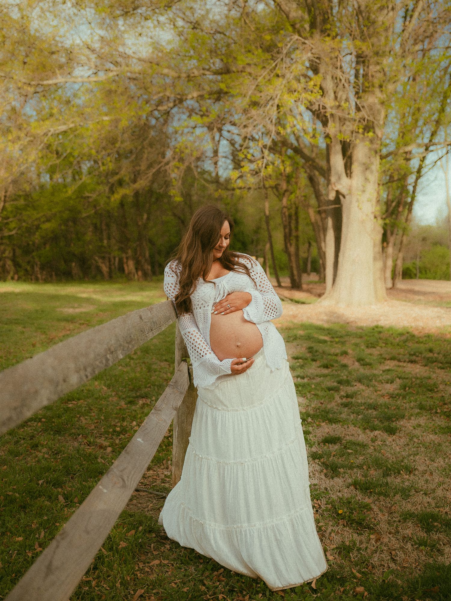 Pregnant woman in white lace maxi dress leaning on fence during outdoor maternity photo session.