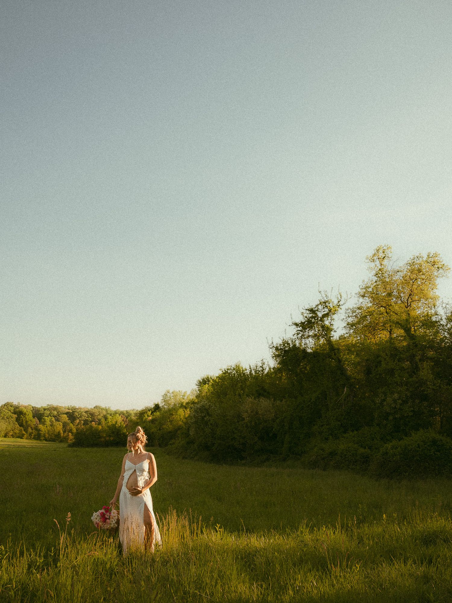 Bride in white gown holding bouquet stands in sunlit green meadow with trees under clear blue sky.