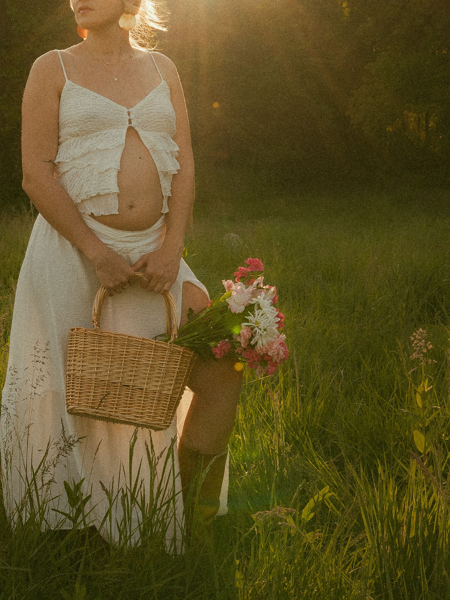 Pregnant woman in white crop top and skirt holding a wicker basket with pink flowers in a sunlit field.
