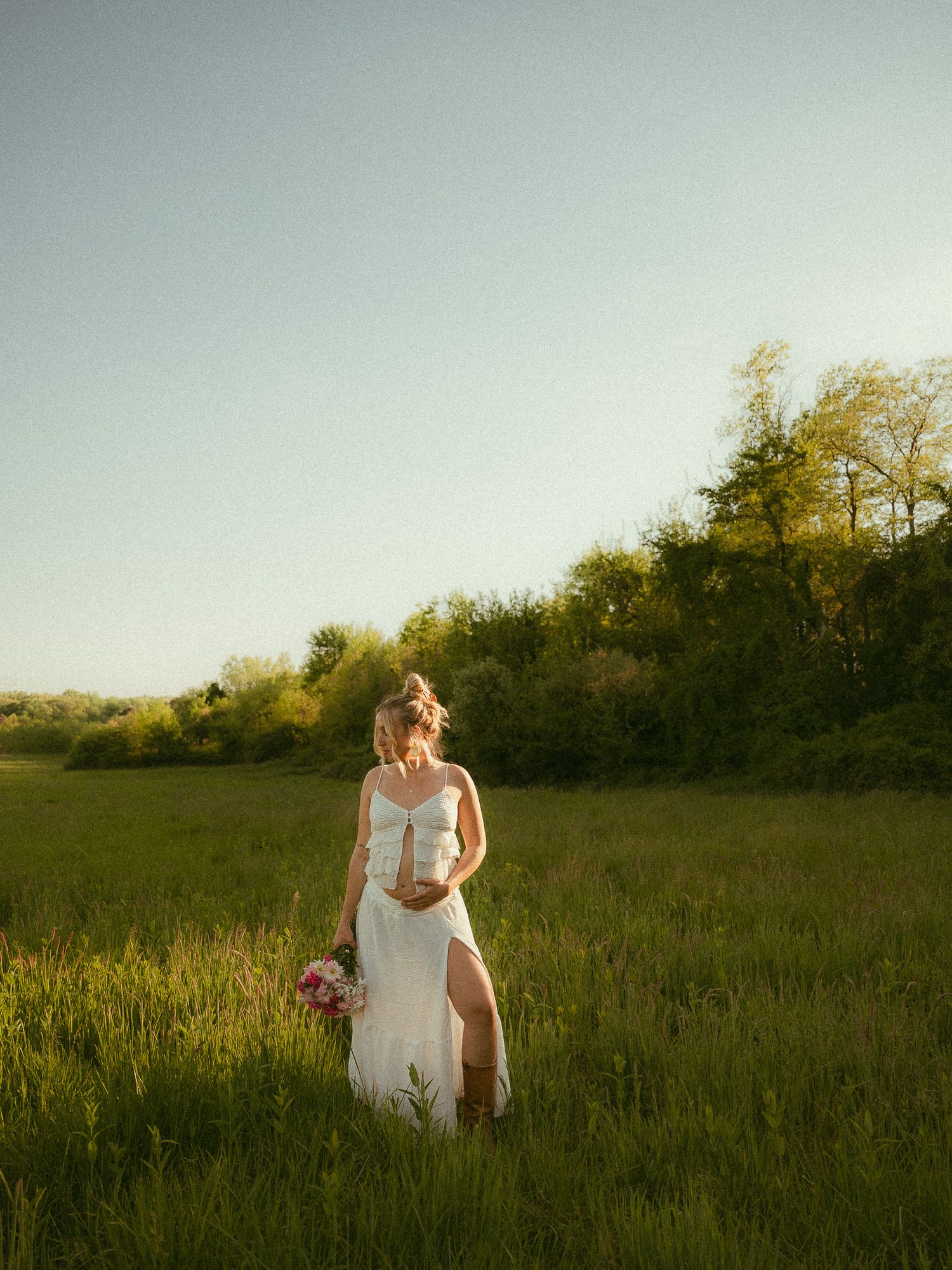 Bride in white gown holding pink bouquet standing in sunlit green meadow at golden hour.