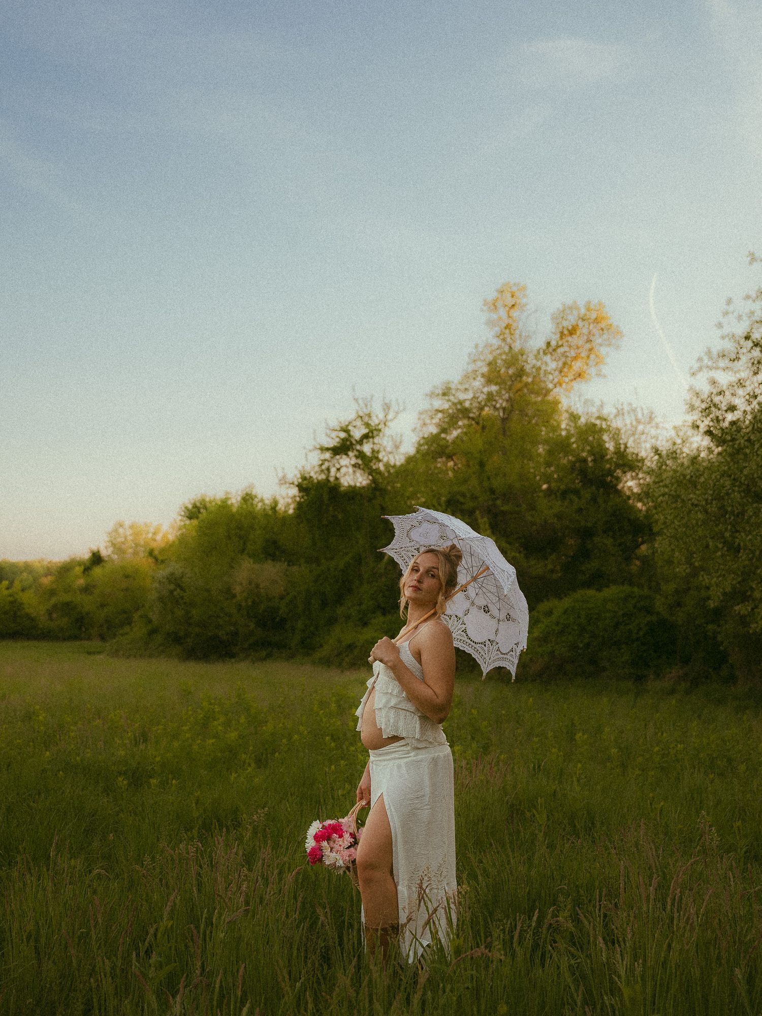 Pregnant woman in white lace dress holding parasol and pink flowers in a green meadow at golden hour.