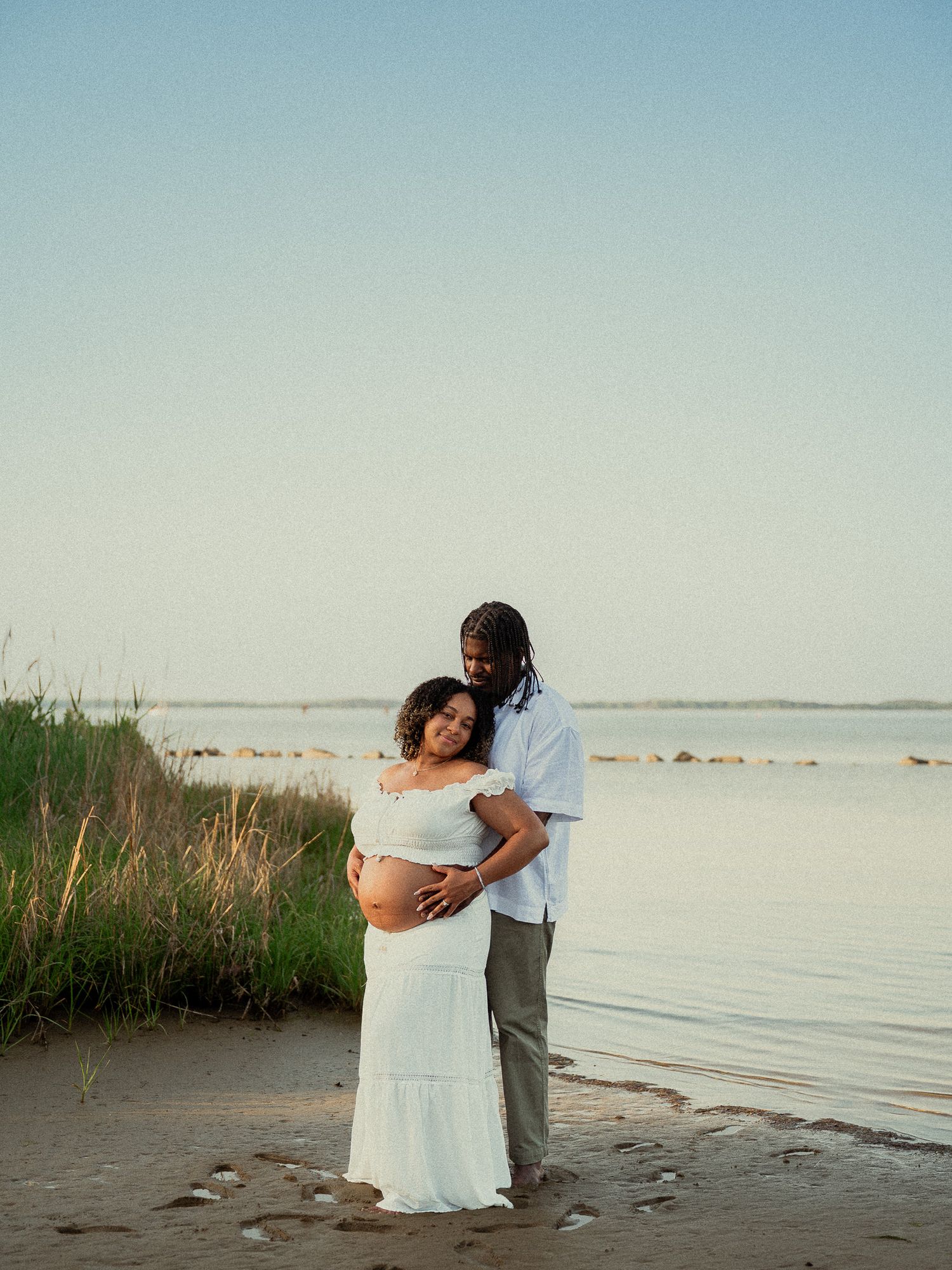 Couple embracing on beach at sunset, pregnant woman in white dress with partner holding her belly.