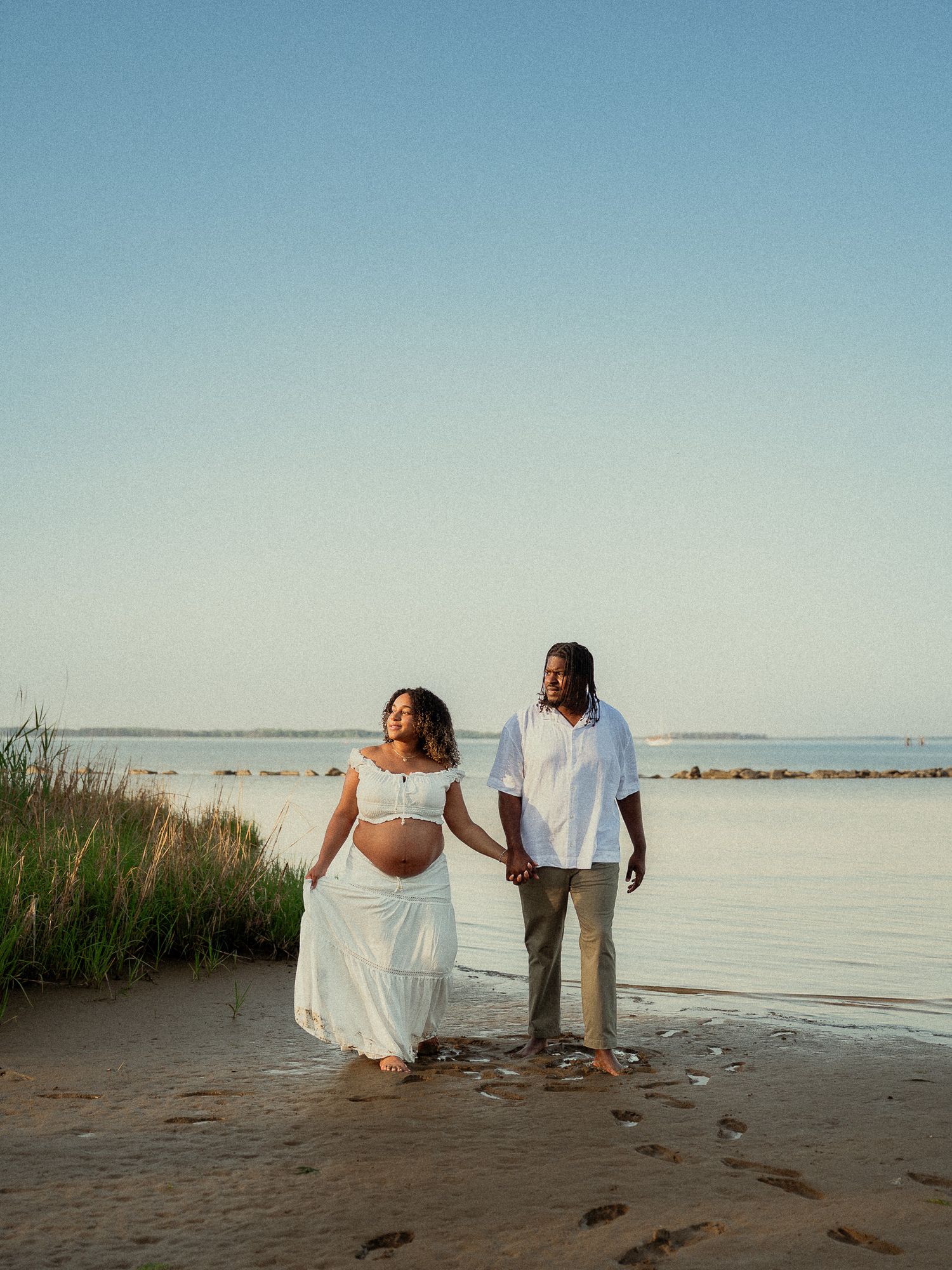 Expecting couple in white outfits hold hands on sandy beach shore with calm water at golden hour.