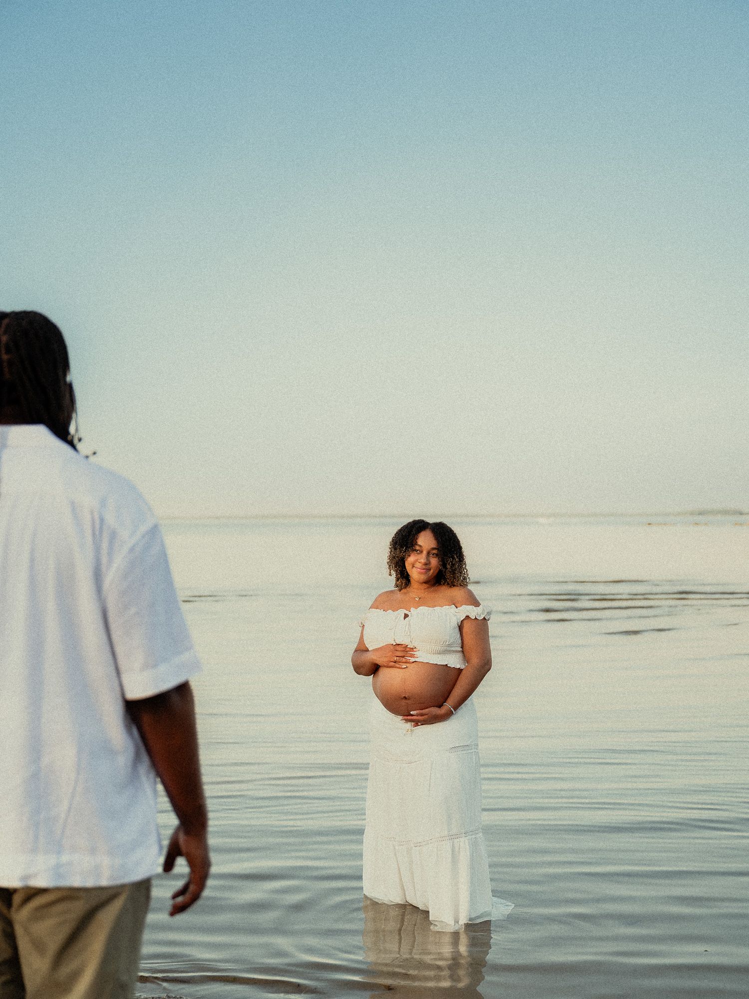 Pregnant woman in white off-shoulder dress stands in shallow ocean water, smiling and cradling her baby bump.