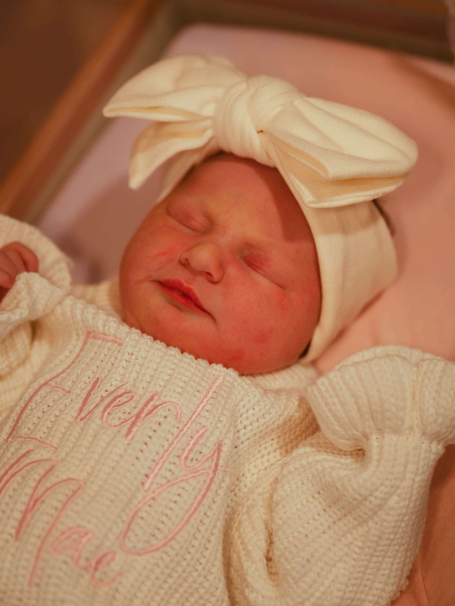Newborn baby girl sleeping peacefully in hospital, wearing white knit outfit and bow headband.