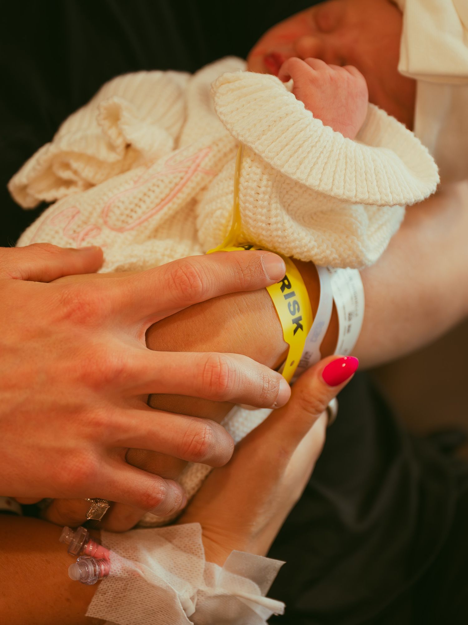 Newborn baby in white knit outfit held by parents, with yellow hospital ID bracelet visible on tiny wrist.