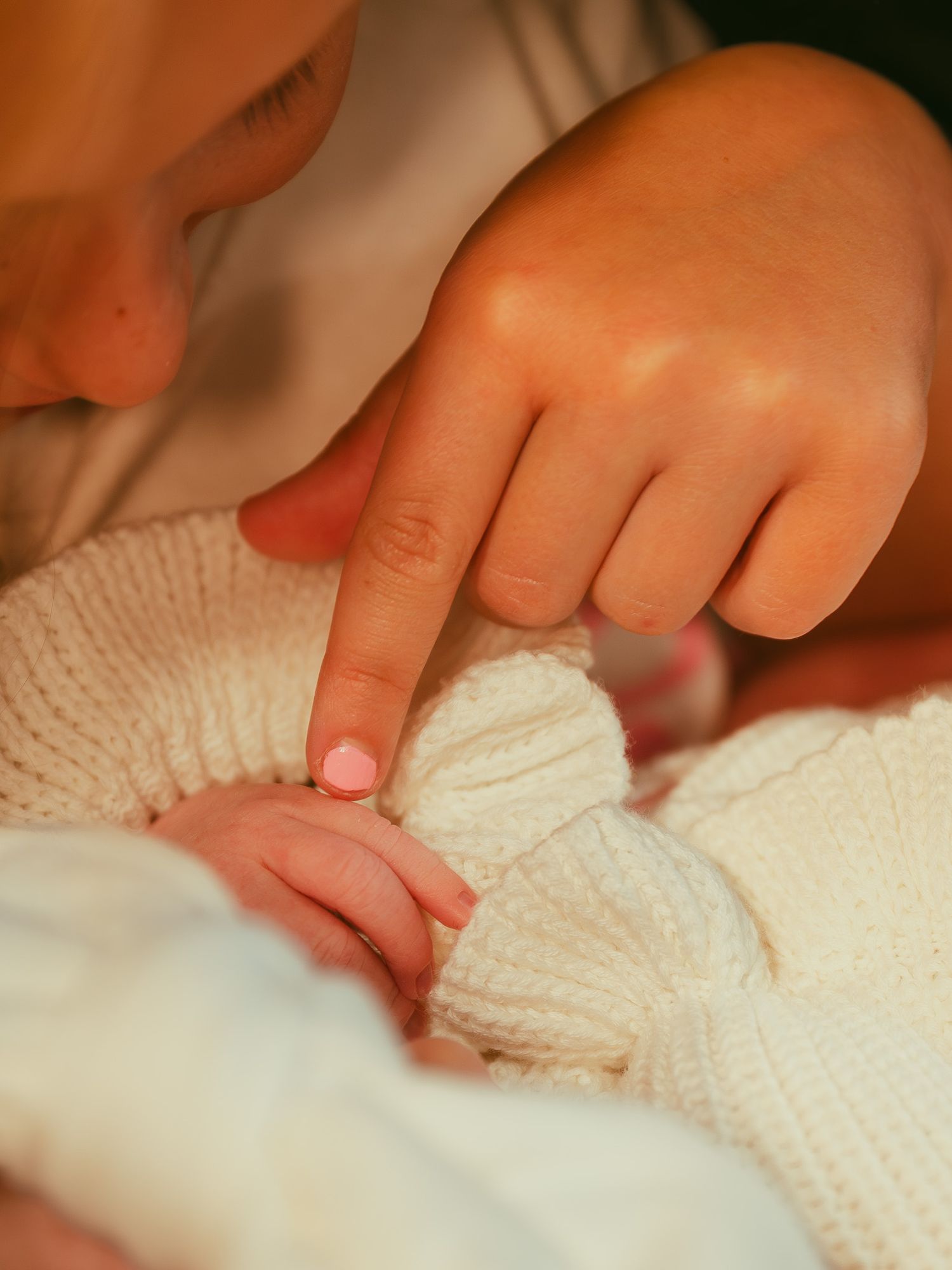 A child gently touches a newborn baby's tiny hand wrapped in a soft white knit blanket.