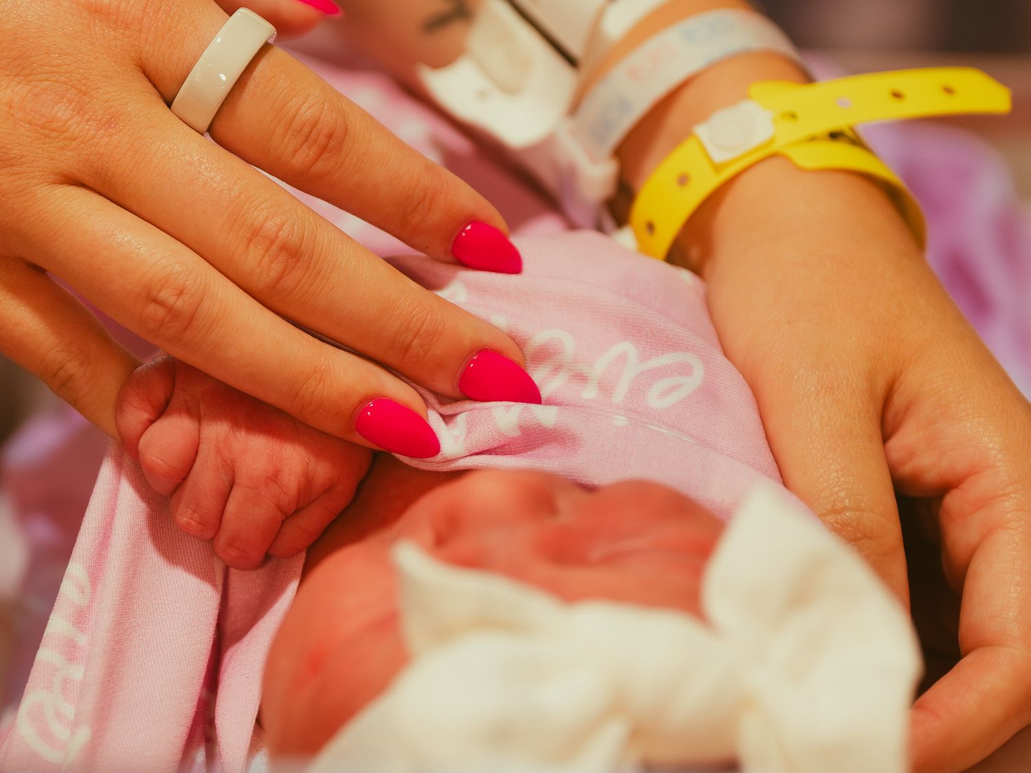 Parents gently hold their newborn baby in pink hat at hospital, mother with red nails wearing ID bracelet.