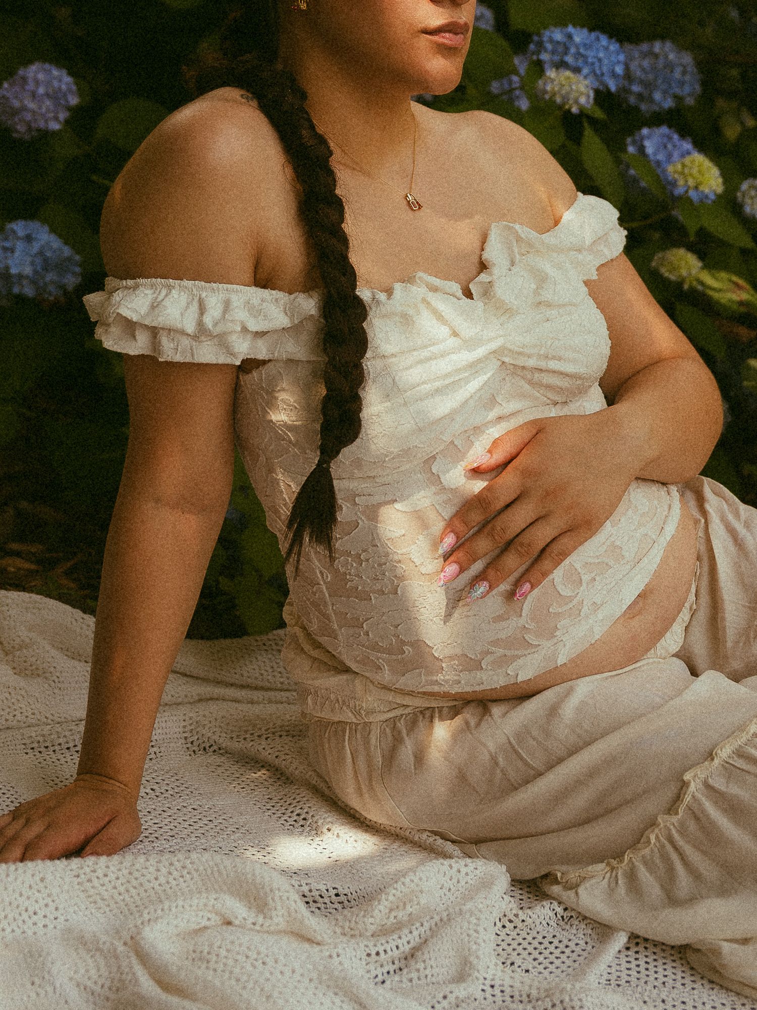 Pregnant woman in white ruffled off-shoulder dress sitting outdoors with hydrangeas, cradling baby bump.