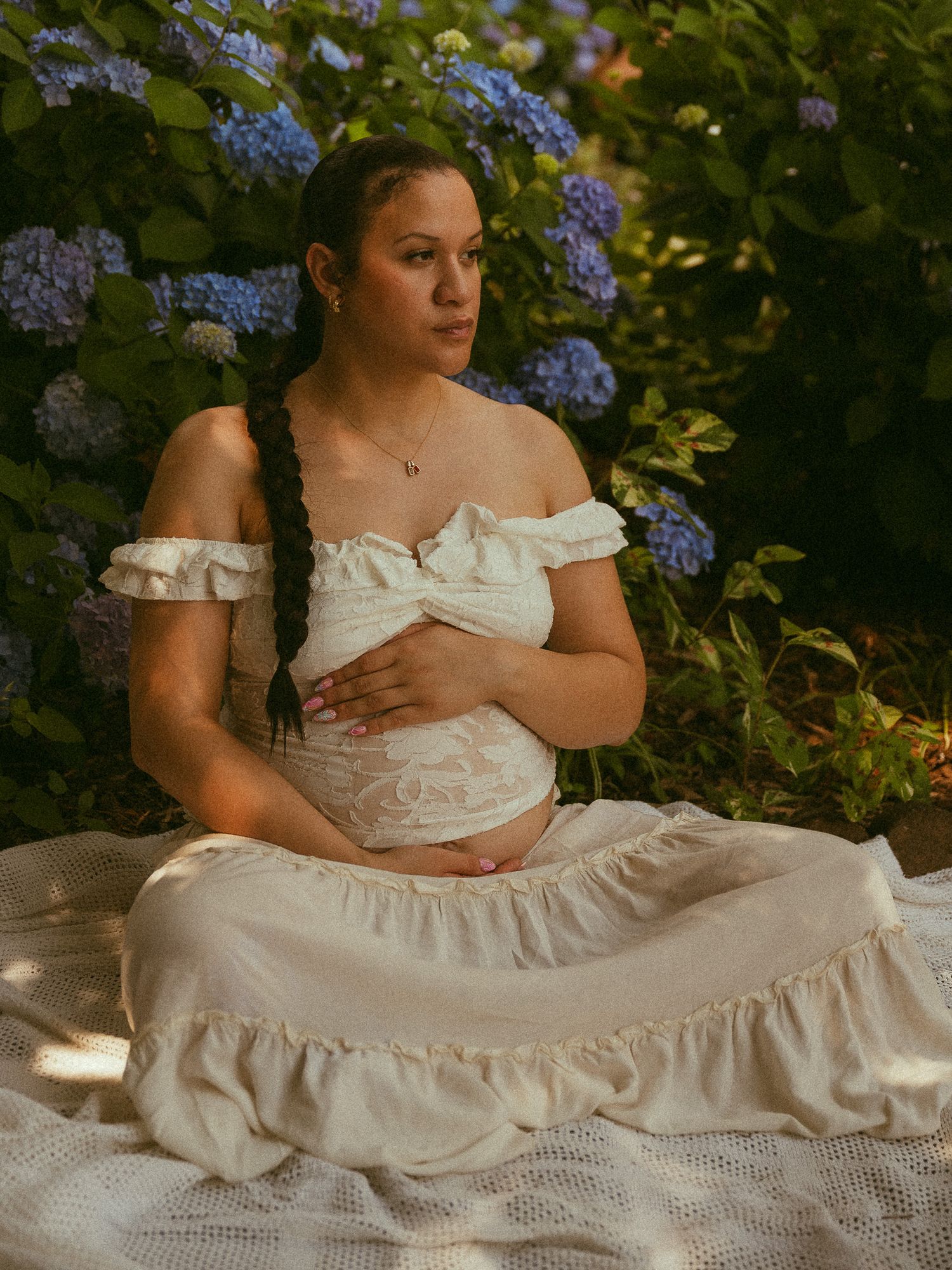 Pregnant woman in white off-shoulder dress sits outdoors among blue hydrangeas, cradling her baby bump.
