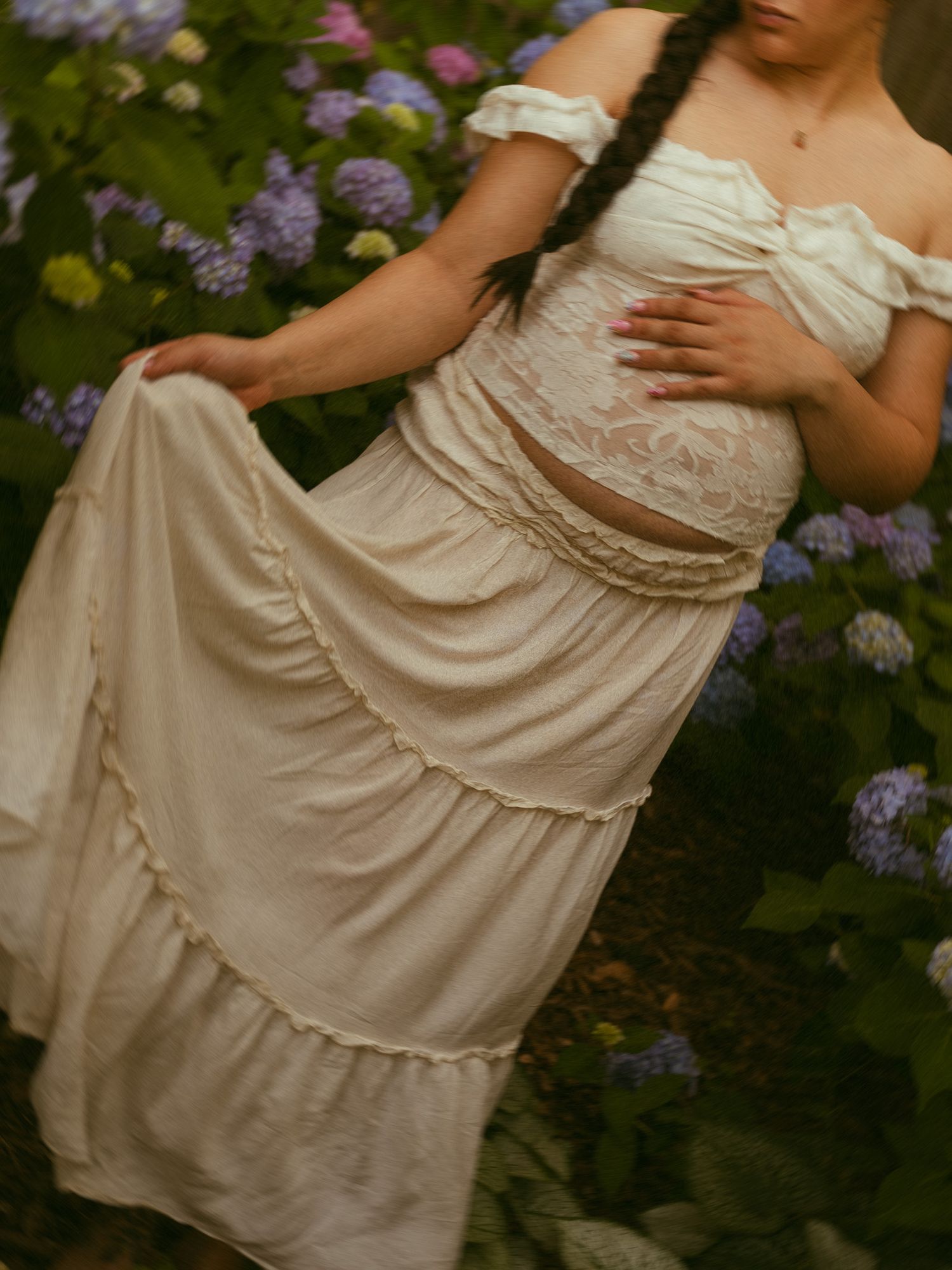 Pregnant woman in white lace crop top and flowing maxi skirt among purple hydrangeas.