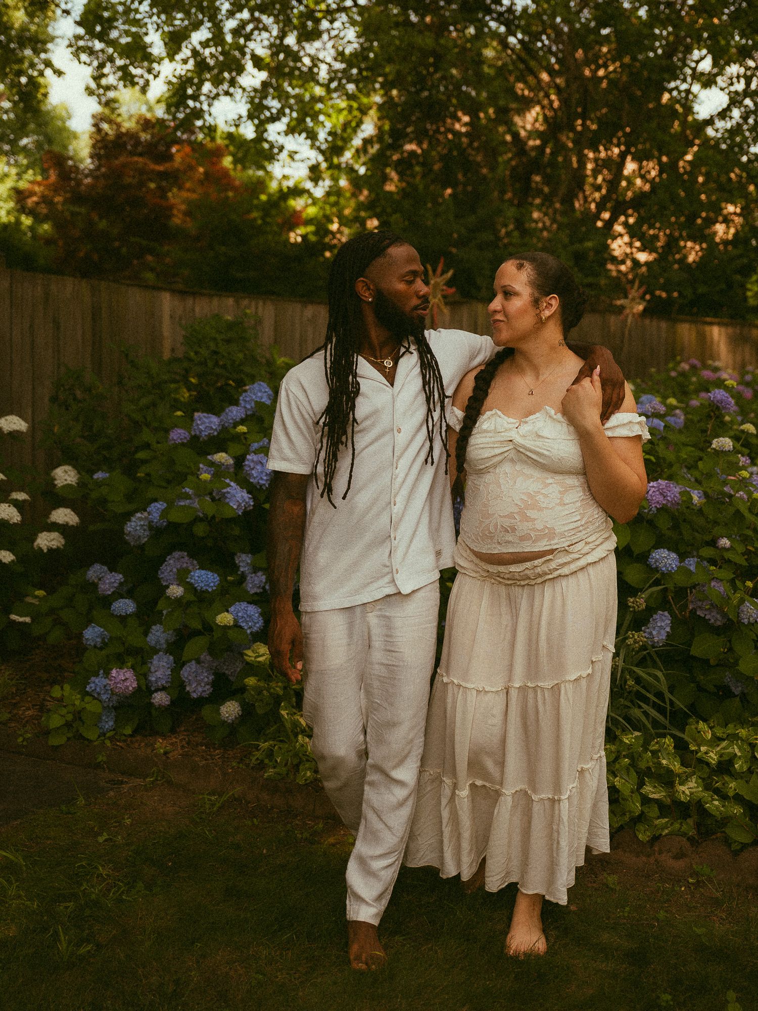 Couple in white outfits standing in a garden with blooming hydrangeas during a maternity photoshoot at golden hour.