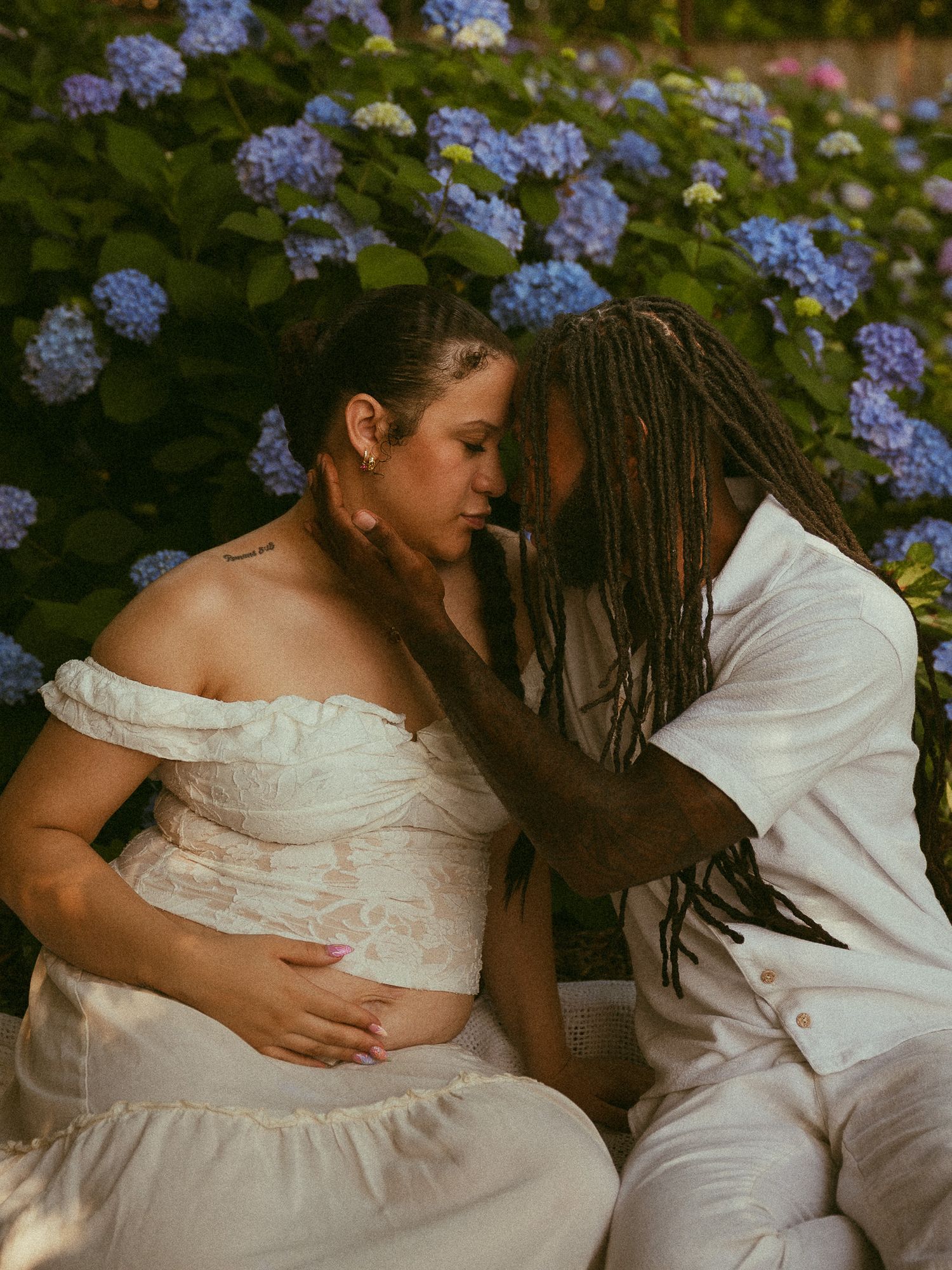 Couple in white outfits sharing tender moment during maternity photoshoot with blue hydrangeas in background.