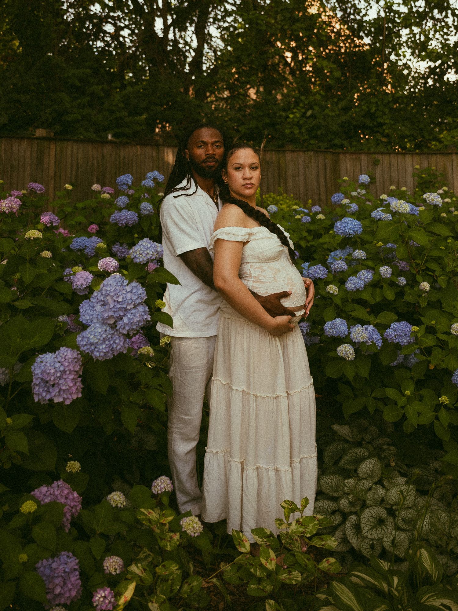 Couple poses in blooming hydrangea garden, woman in white maternity dress cradling baby bump, man embracing her from behind.