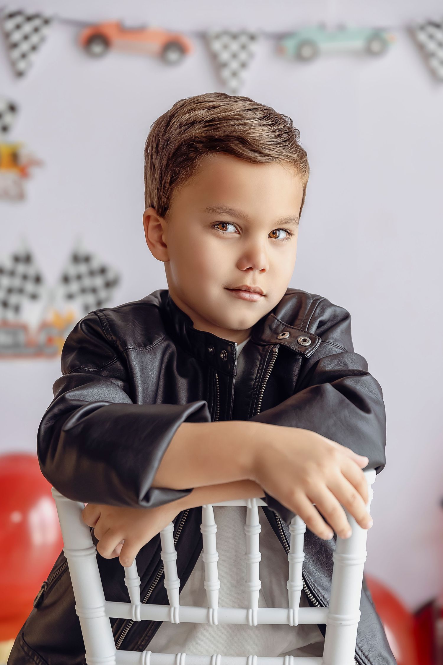 Nino de 5 anos mirando a la camara con una pequena sonrisa y un look cool sentando en una silla la sesion fotografica es de carros