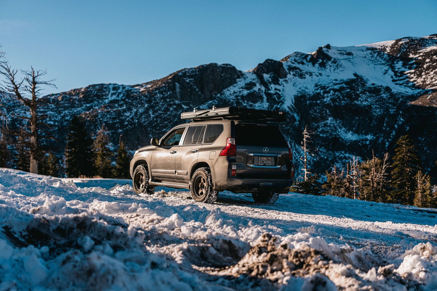A brown SUV drives through a snowy mountain landscape with dramatic snow-capped peaks in the background.