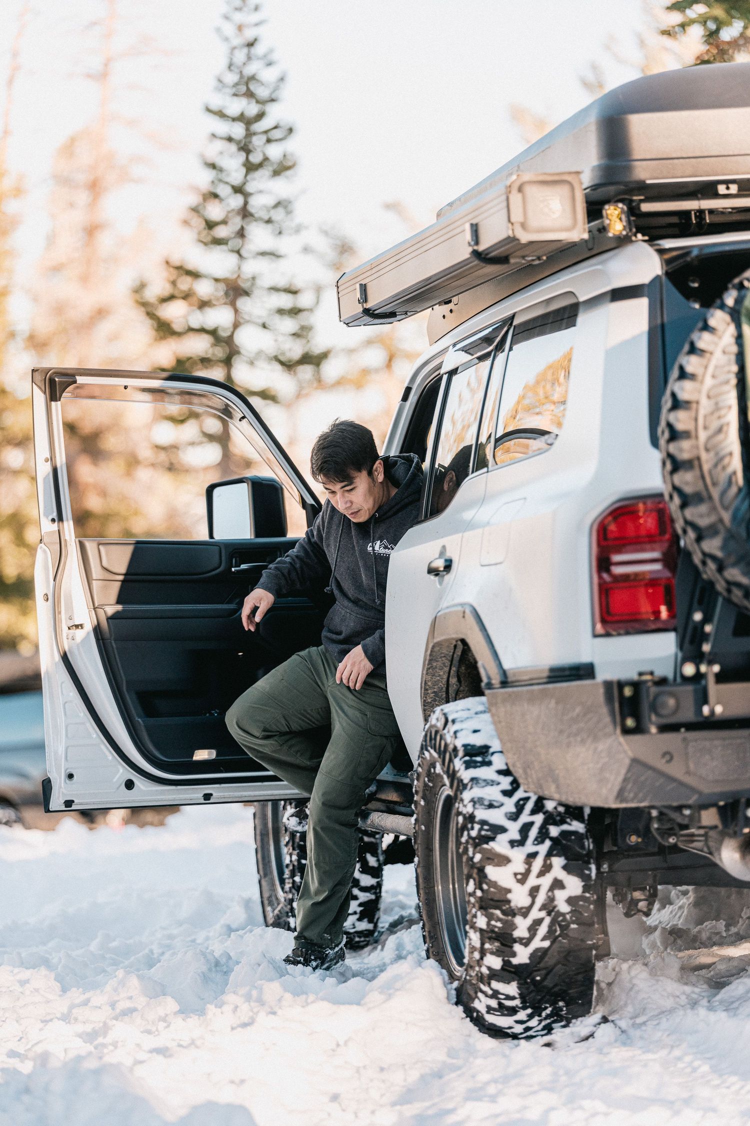 Person in winter attire sits in the open trunk of a car parked in snowy winter conditions.